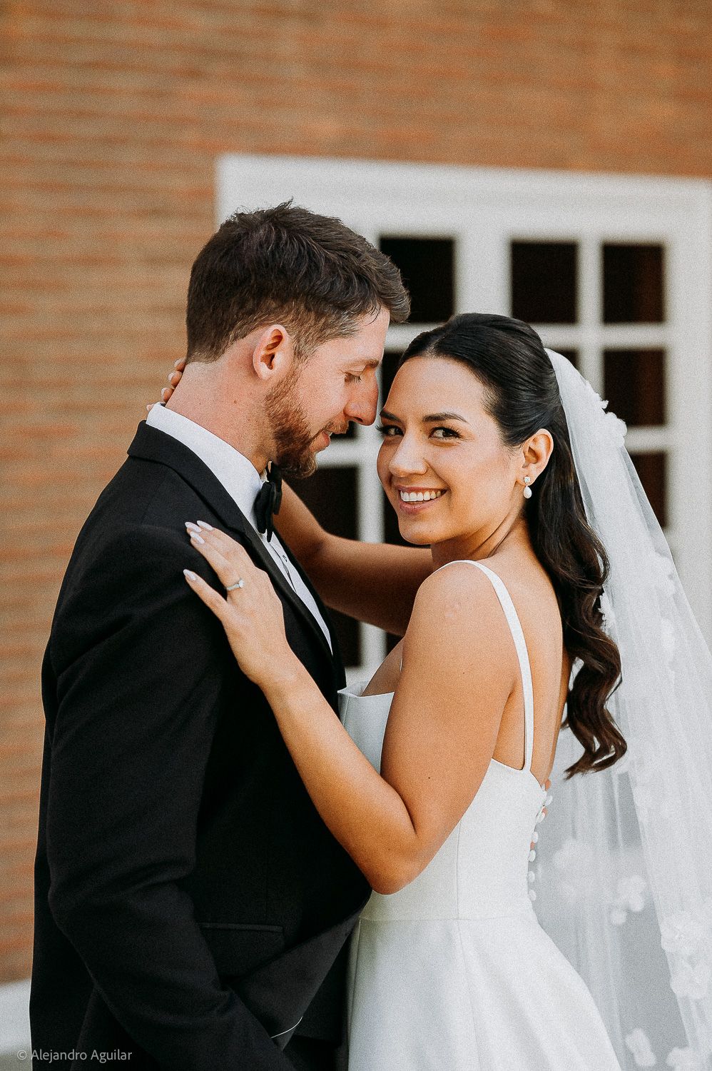 Bride in white gown and veil smiles at camera while groom in black tuxedo leans in close outdoors.