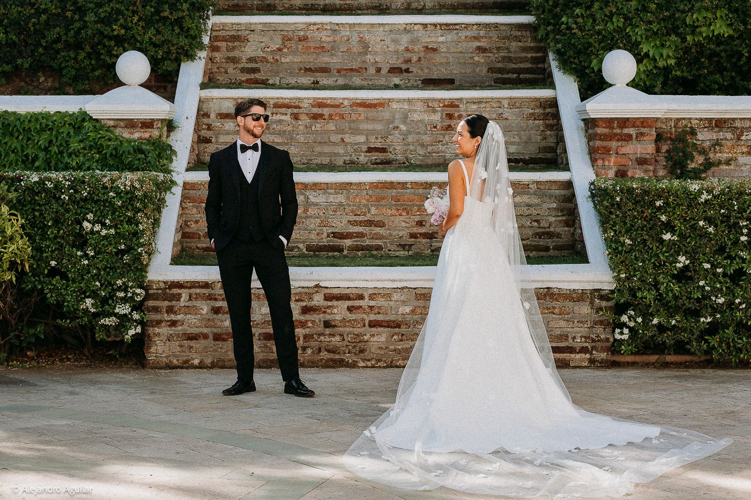 Bride in elegant white gown with veil and groom in black tuxedo pose in a garden with brick steps.