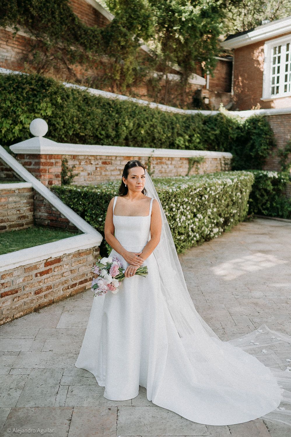 Bride in white gown with veil holds pink bouquet outdoors at elegant brick garden venue.