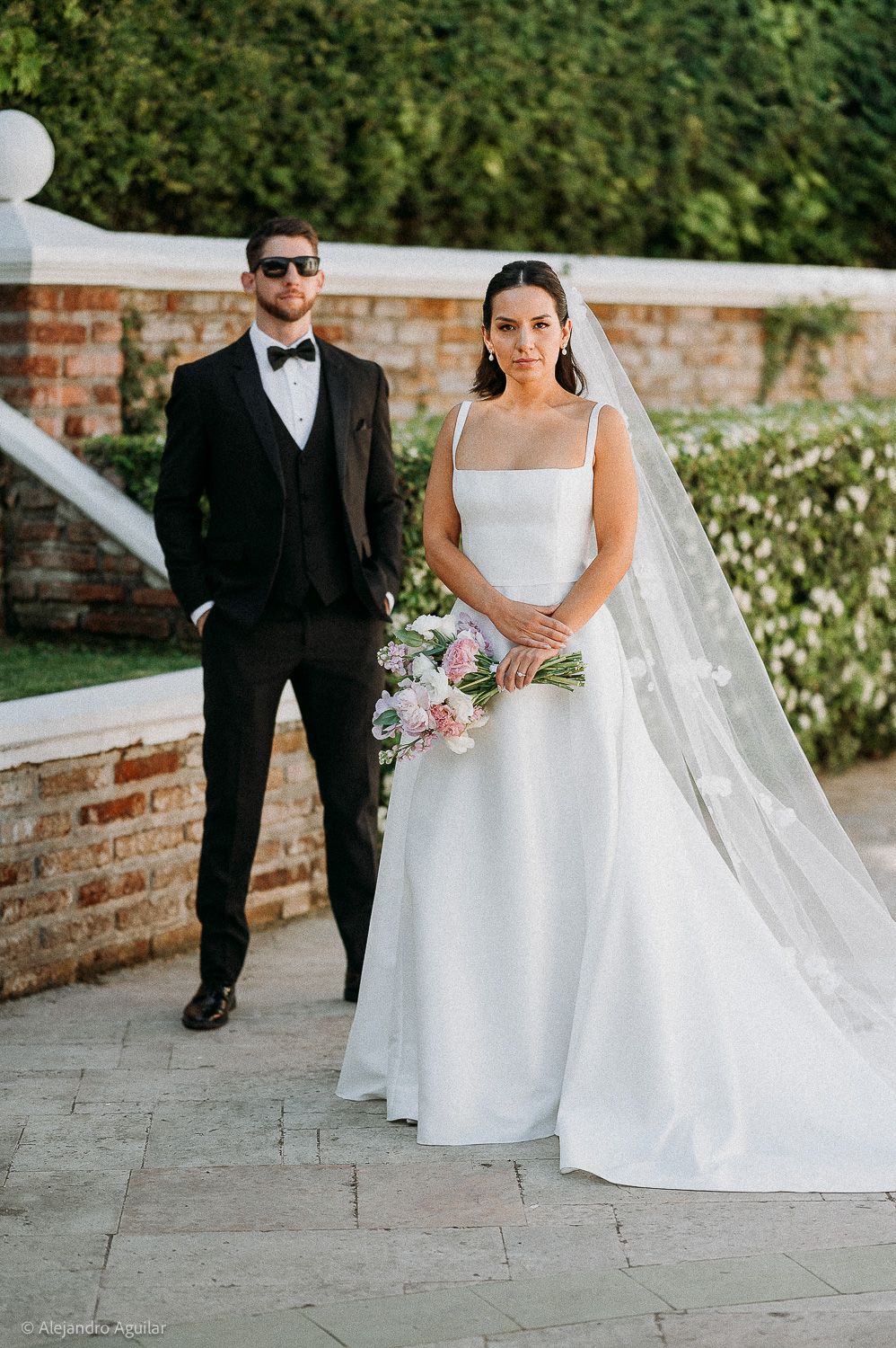 Bride in white gown with veil holds bouquet beside groom in black tuxedo at outdoor wedding venue.