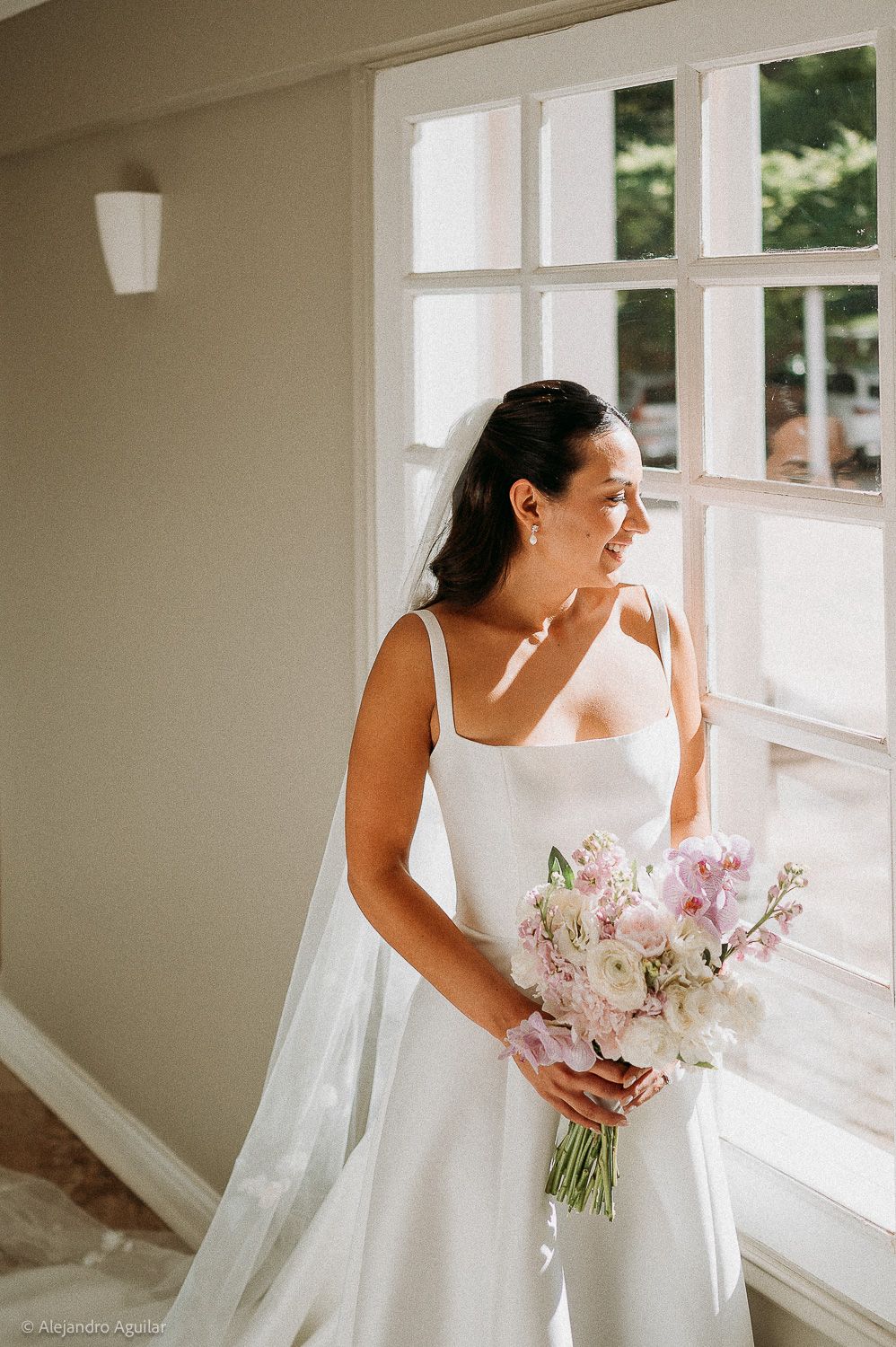 Bride in white gown holding pink and white bouquet gazes out window on wedding day.
