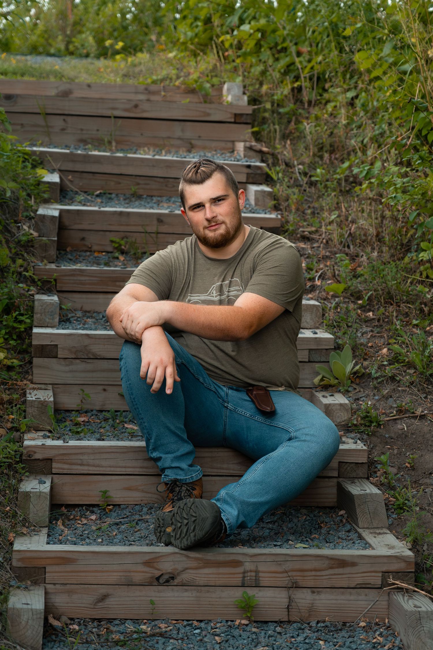 Young man in green t-shirt and jeans sitting relaxed on rustic wooden stairs surrounded by lush greenery.