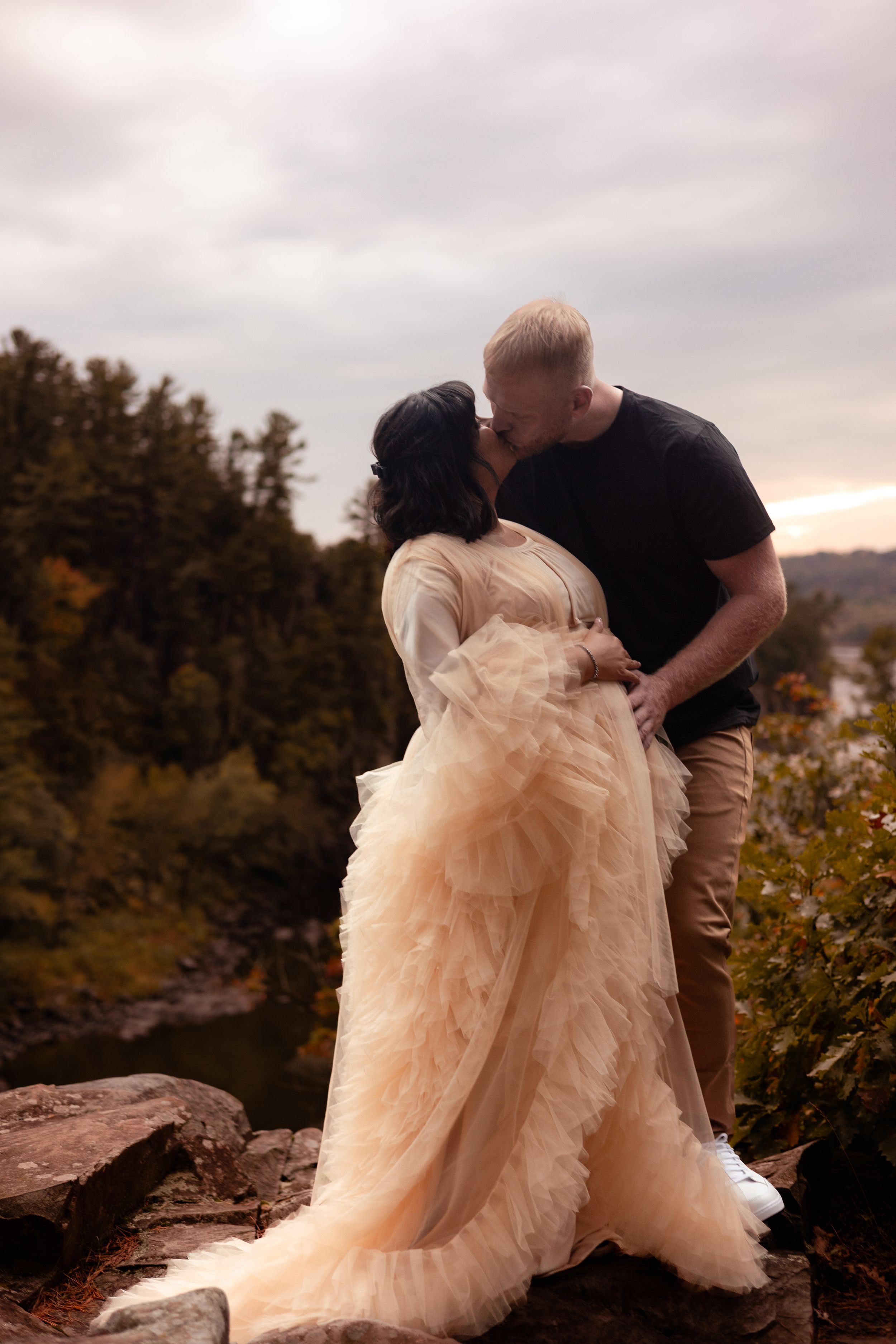 A couple shares a romantic kiss outdoors, woman in a flowing champagne tulle gown, surrounded by lush trees and a river.