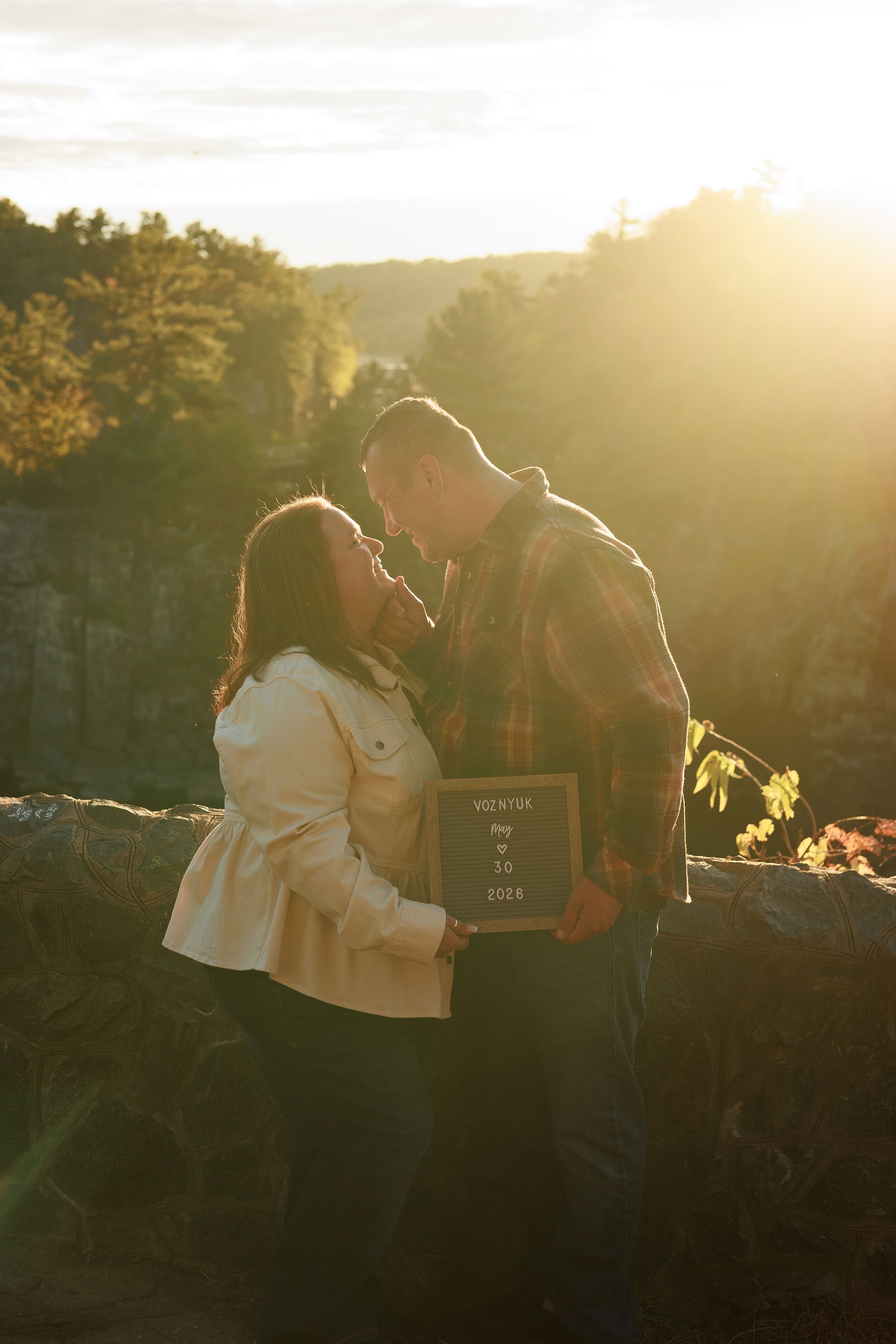 Couple sharing a romantic moment at golden hour outdoors, holding a sign, surrounded by autumn foliage and warm sunlight.