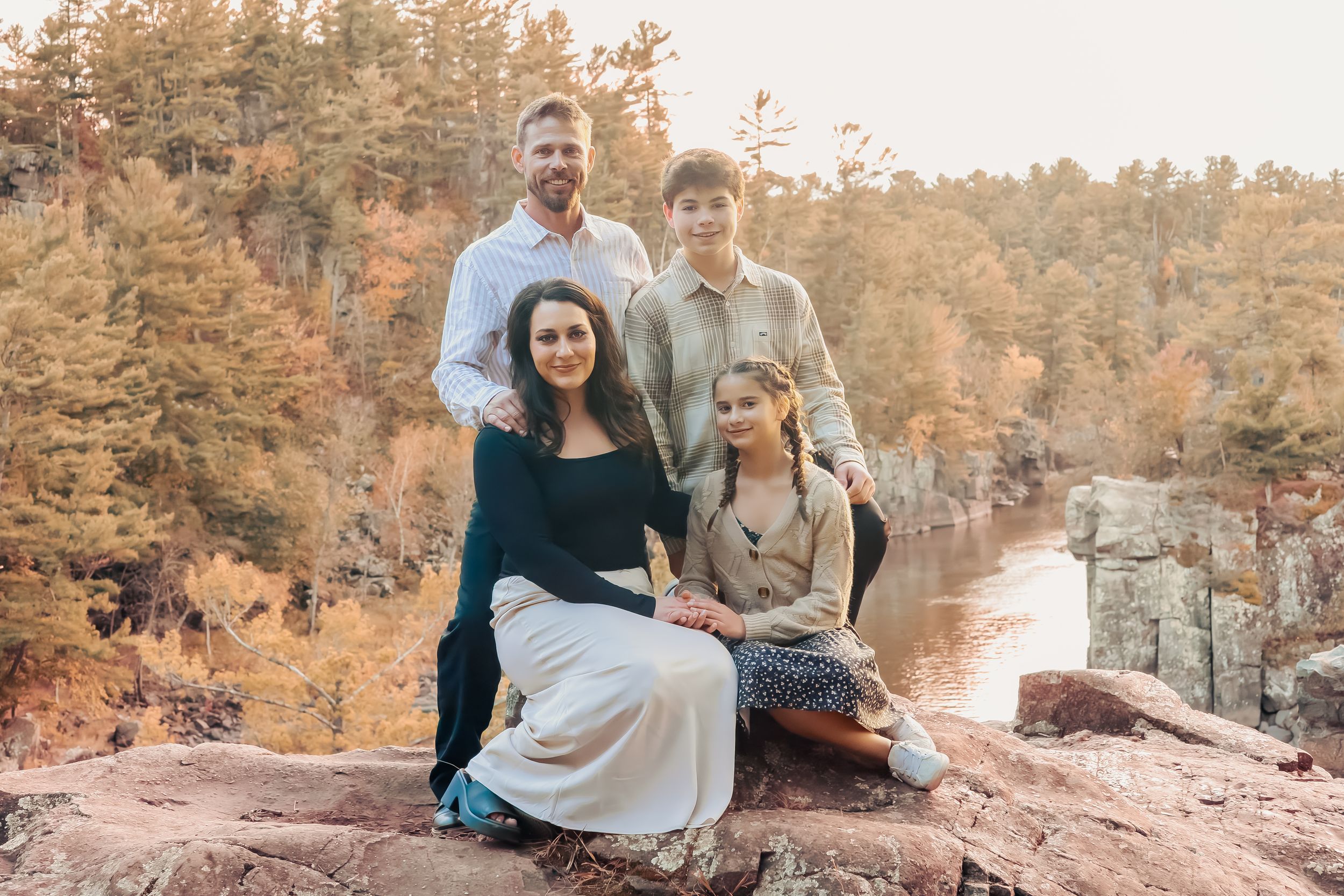 Family of four posing on rocky cliff overlooking river surrounded by autumn forest trees.