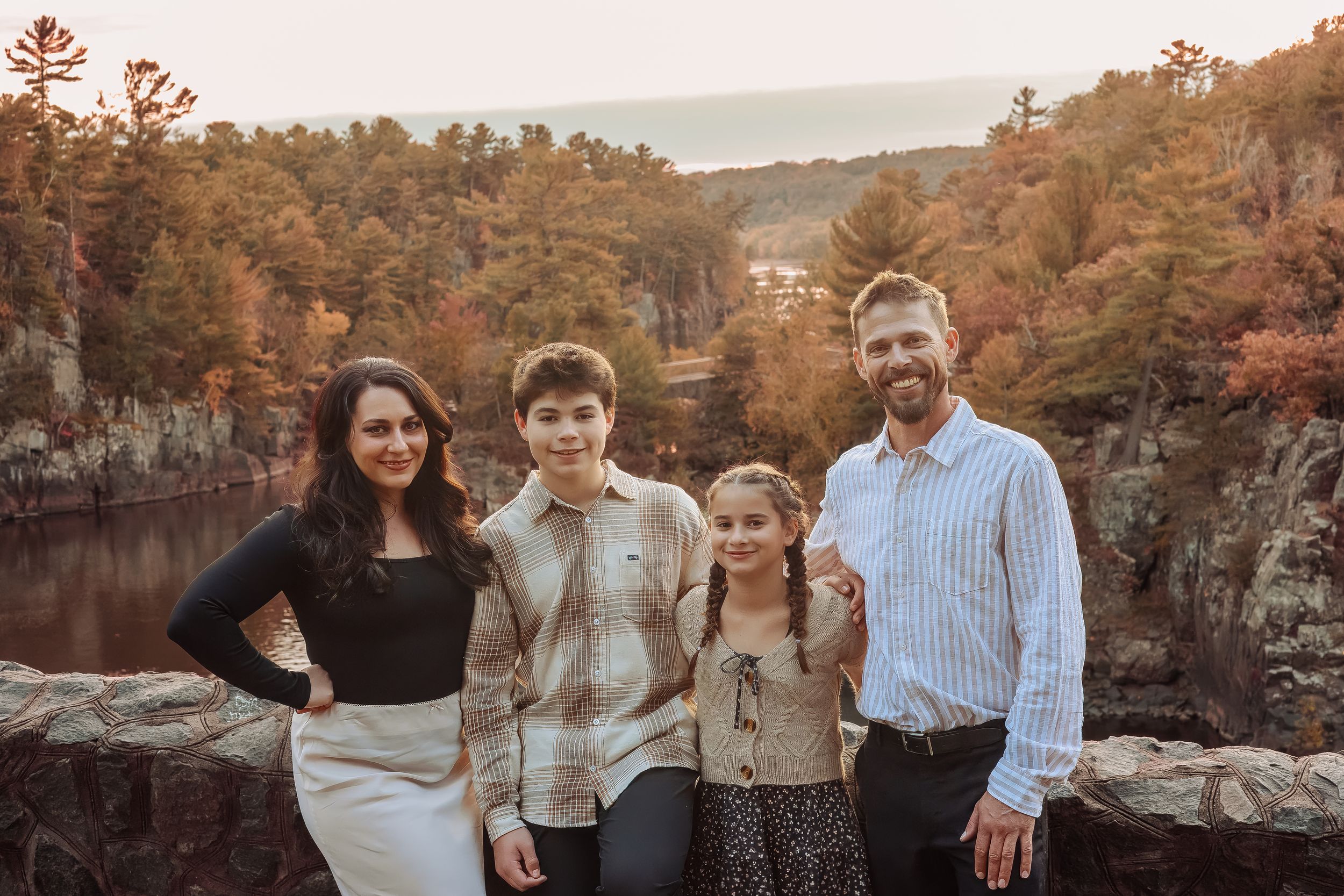 Family of four smiling together on a stone overlook surrounded by colorful autumn foliage and a scenic river below.