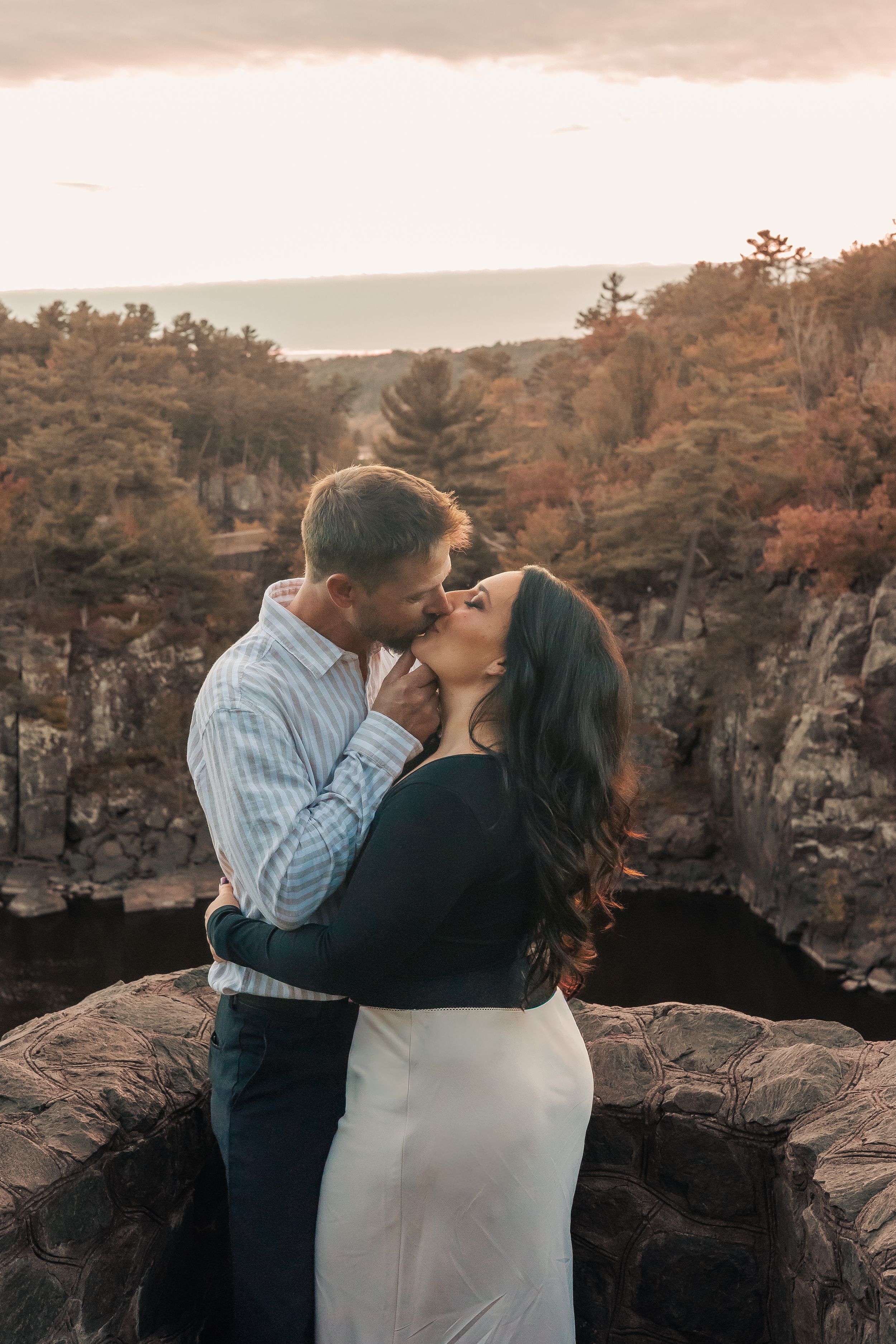 A couple shares a romantic kiss on rocky cliffs surrounded by autumn foliage and a scenic overlook at sunset.