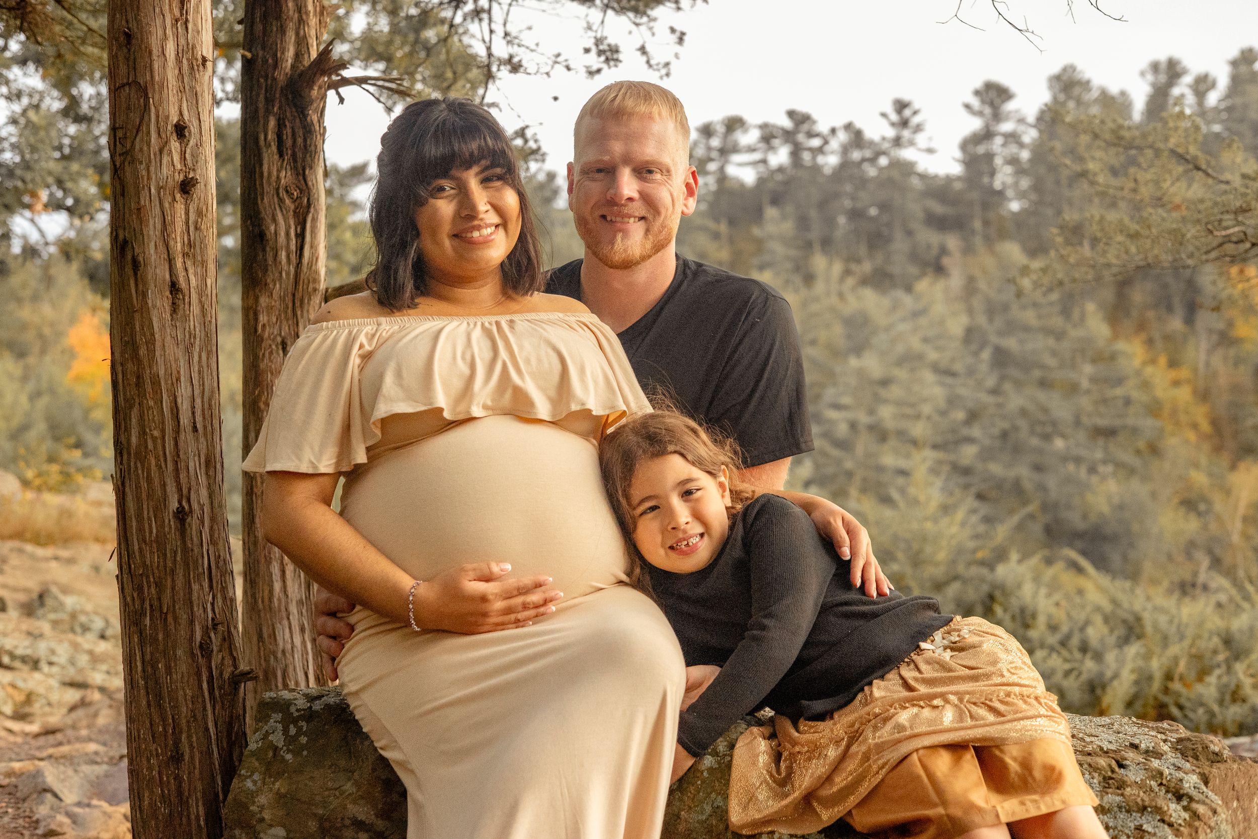 Pregnant woman in cream dress with husband and daughter posing by tree in outdoor fall maternity photo session.