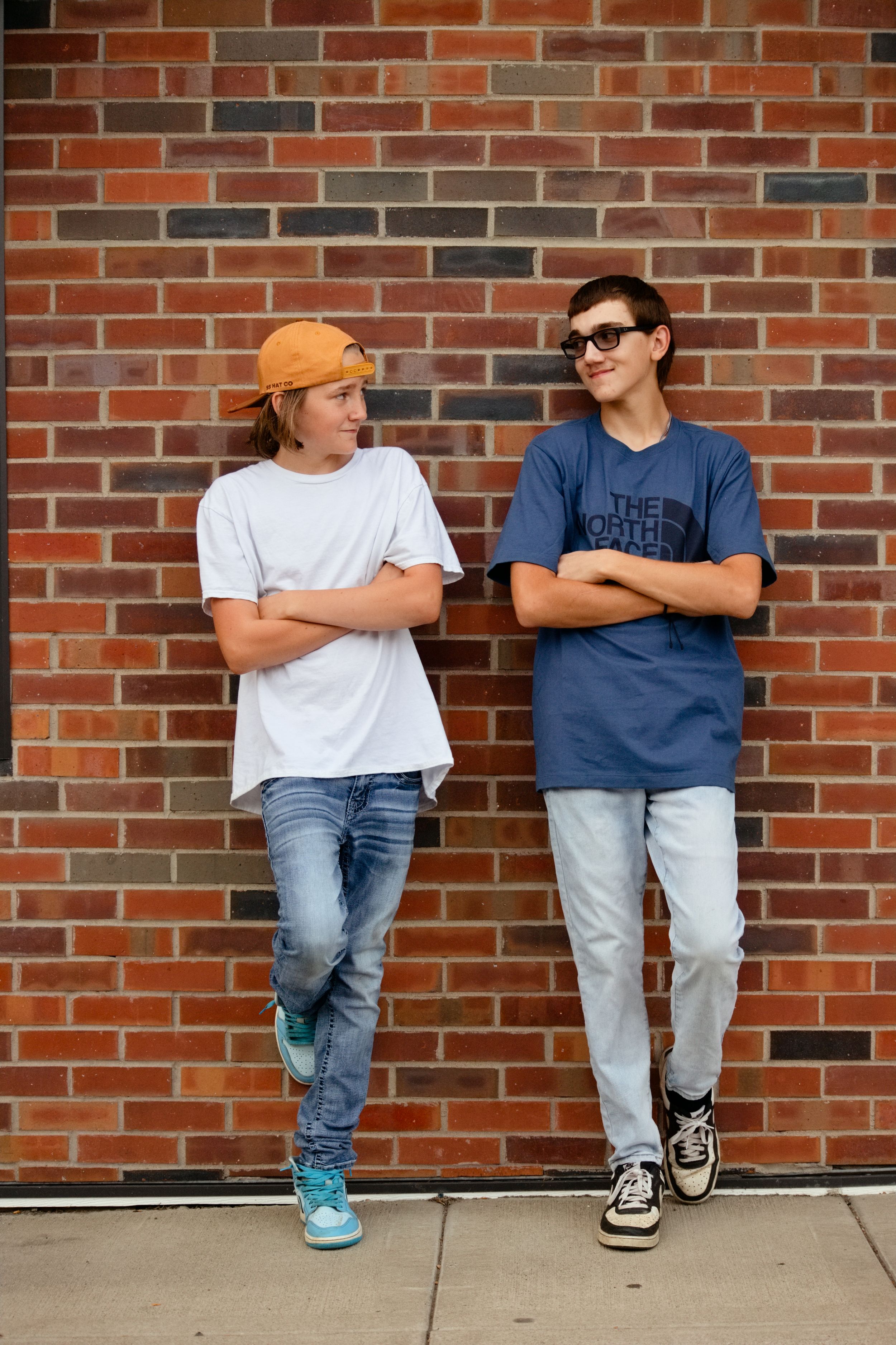 Two teenage boys leaning against a brick wall, one in an orange cap and white tee, the other in a North Face blue shirt.