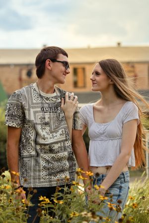 Young couple smiling at each other in a flower garden, man in Guess t-shirt and sunglasses, woman in white crop top.