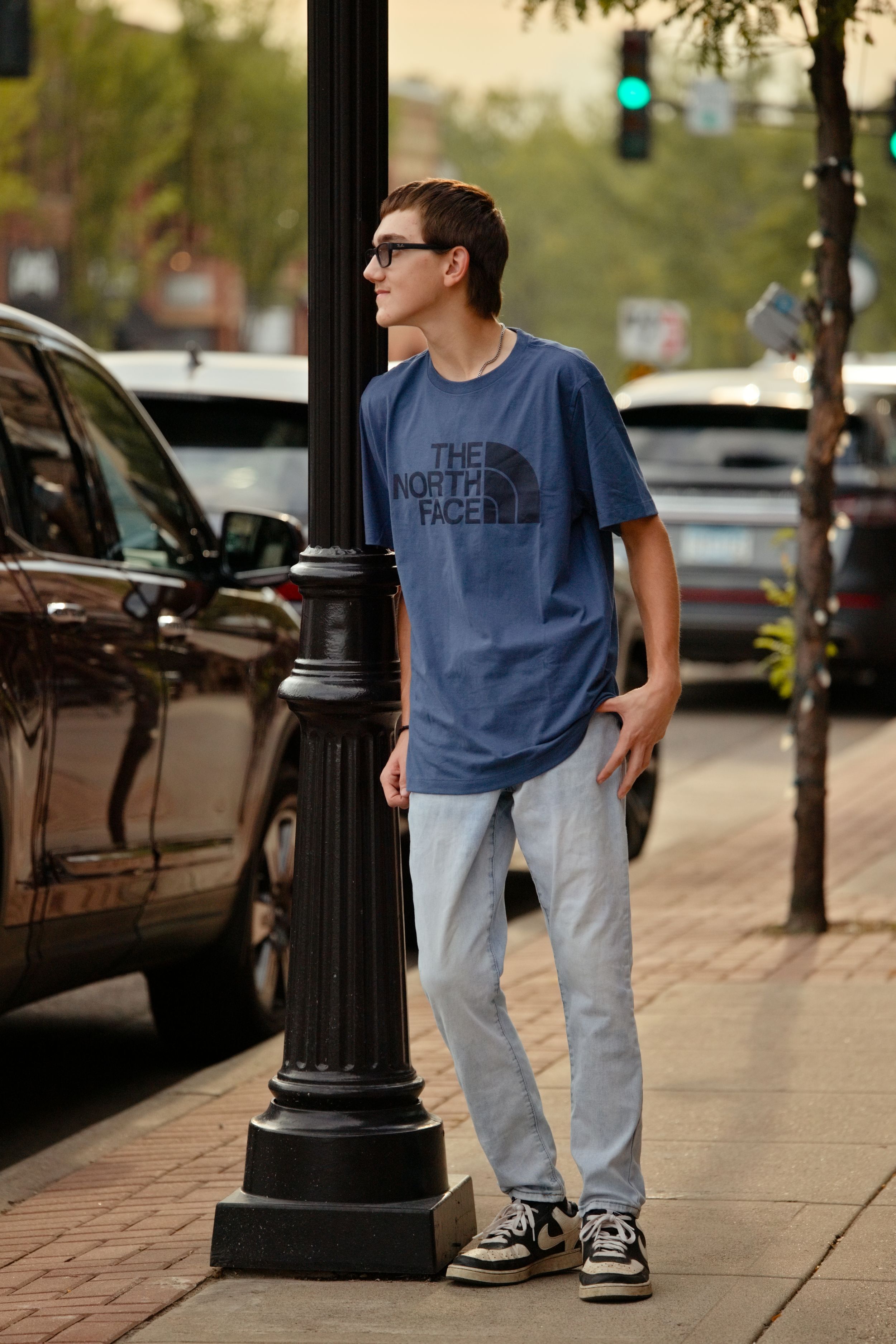 Young man in North Face blue tee and light jeans leaning on street lamp post on urban sidewalk.