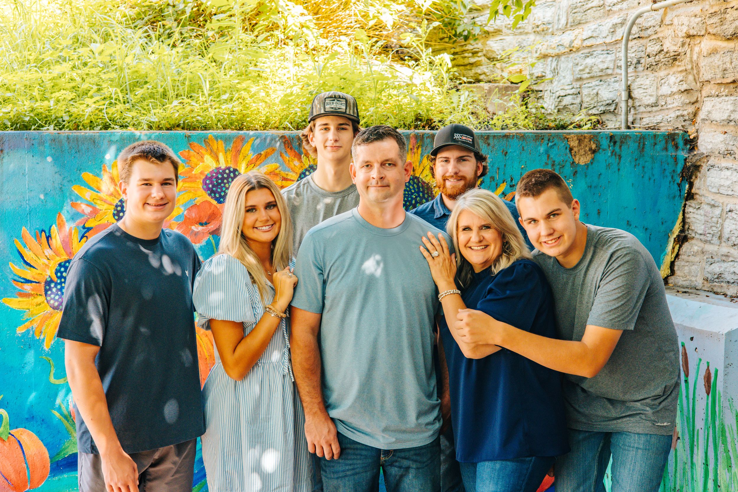 A joyful family of seven poses together in front of a colorful sunflower mural outdoors.