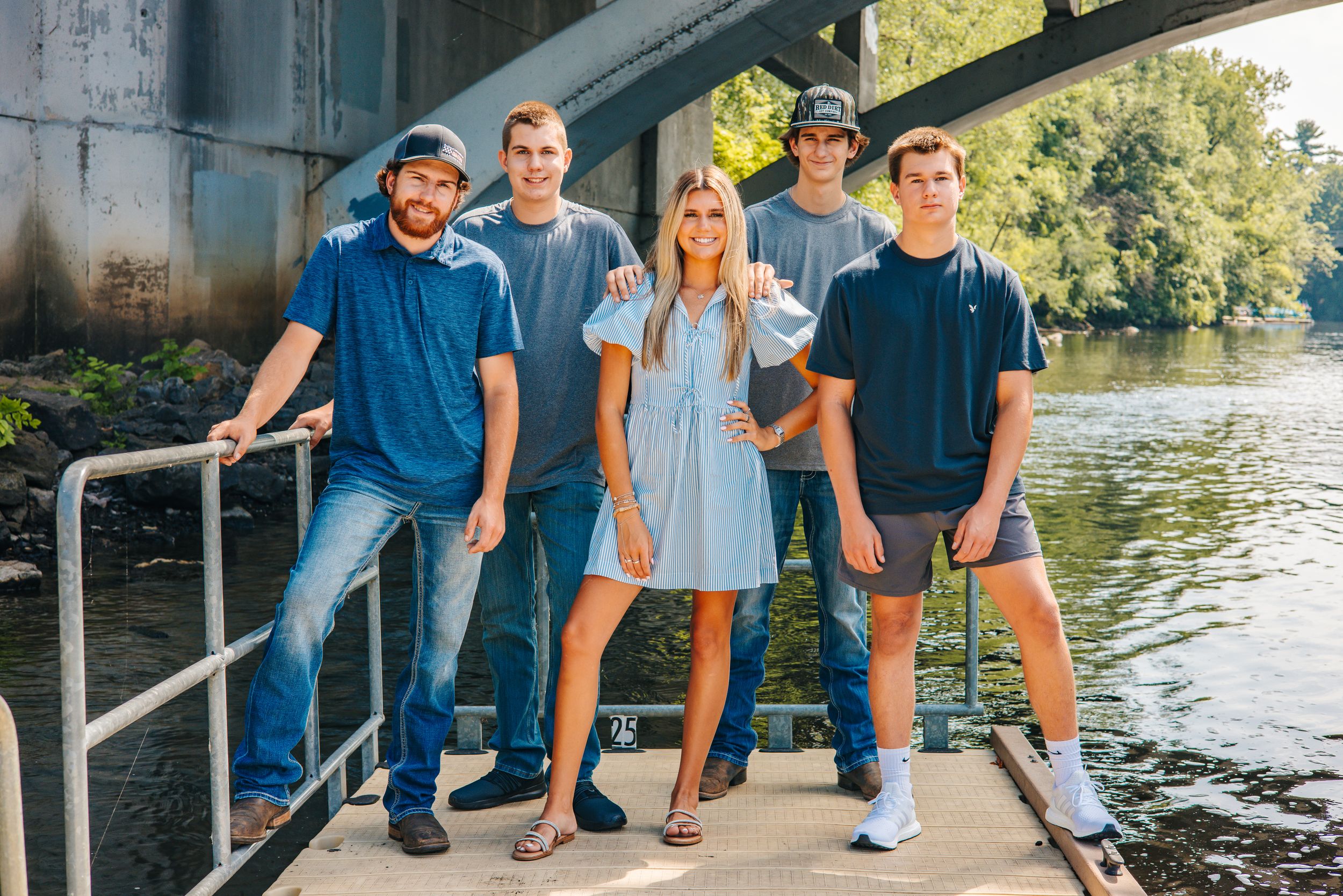 Five people posing together under a bridge near a river on a sunny day, wearing casual blue outfits.