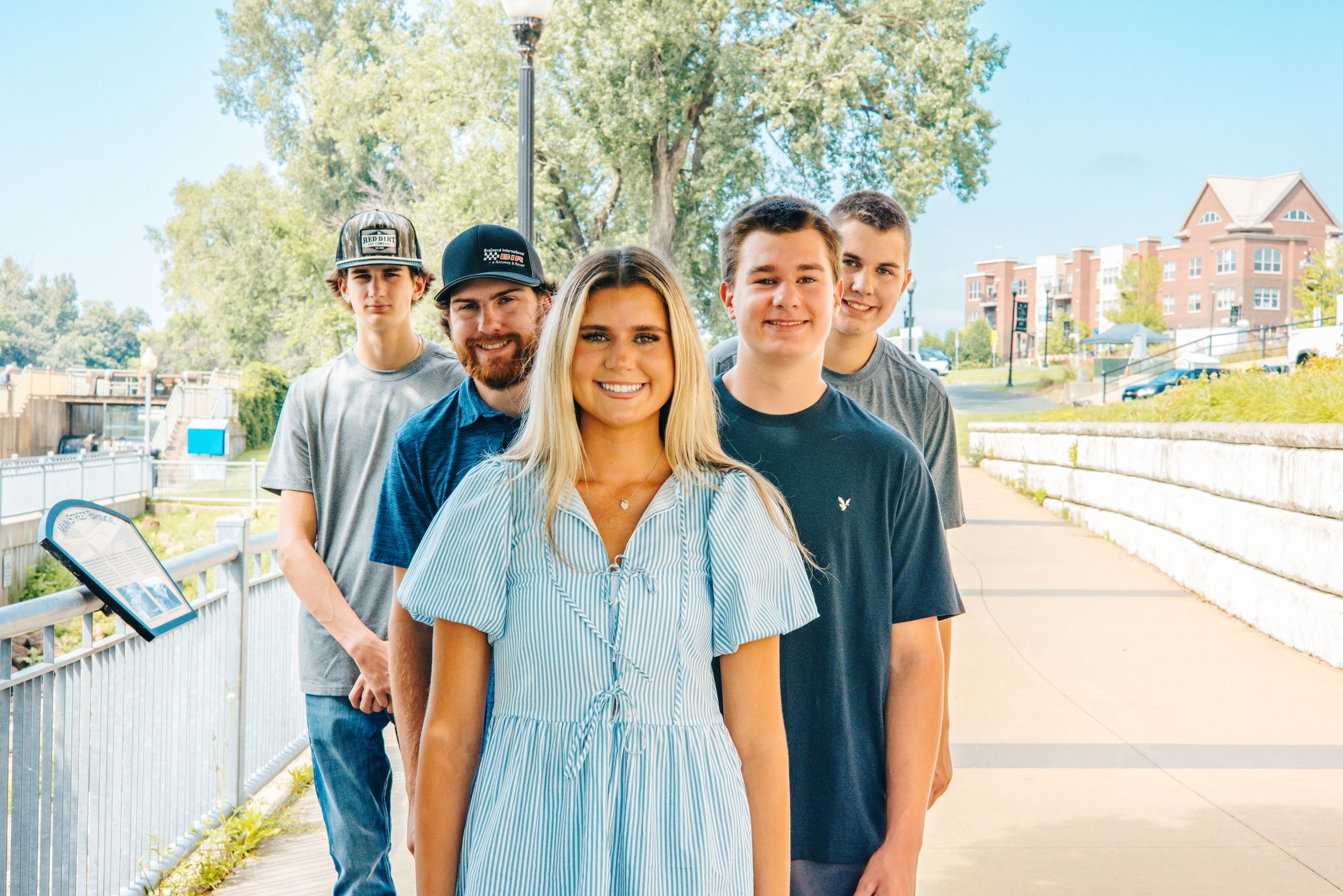 Group of five young adults posing on a sunny sidewalk, smiling outdoors near a brick building.