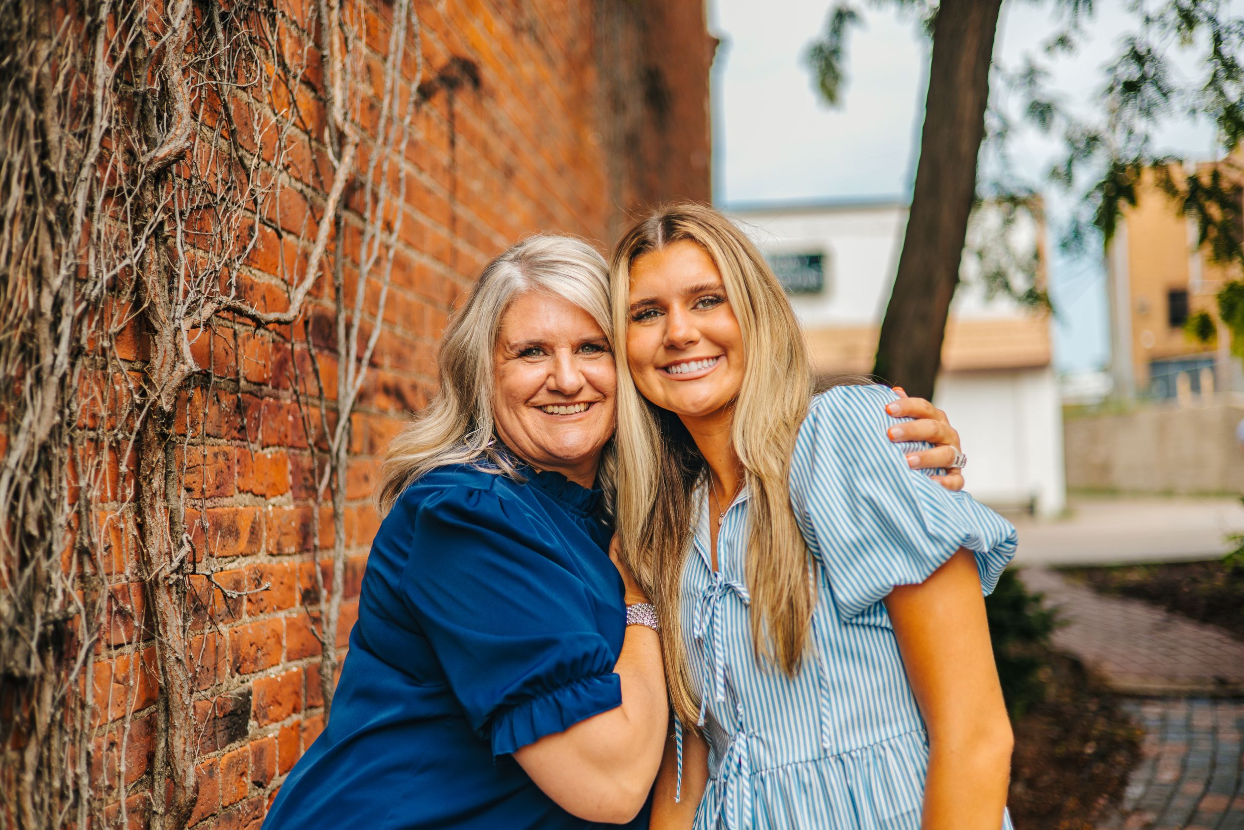 Two women smiling and hugging outdoors near a brick wall, one in blue top and one in light blue dress.