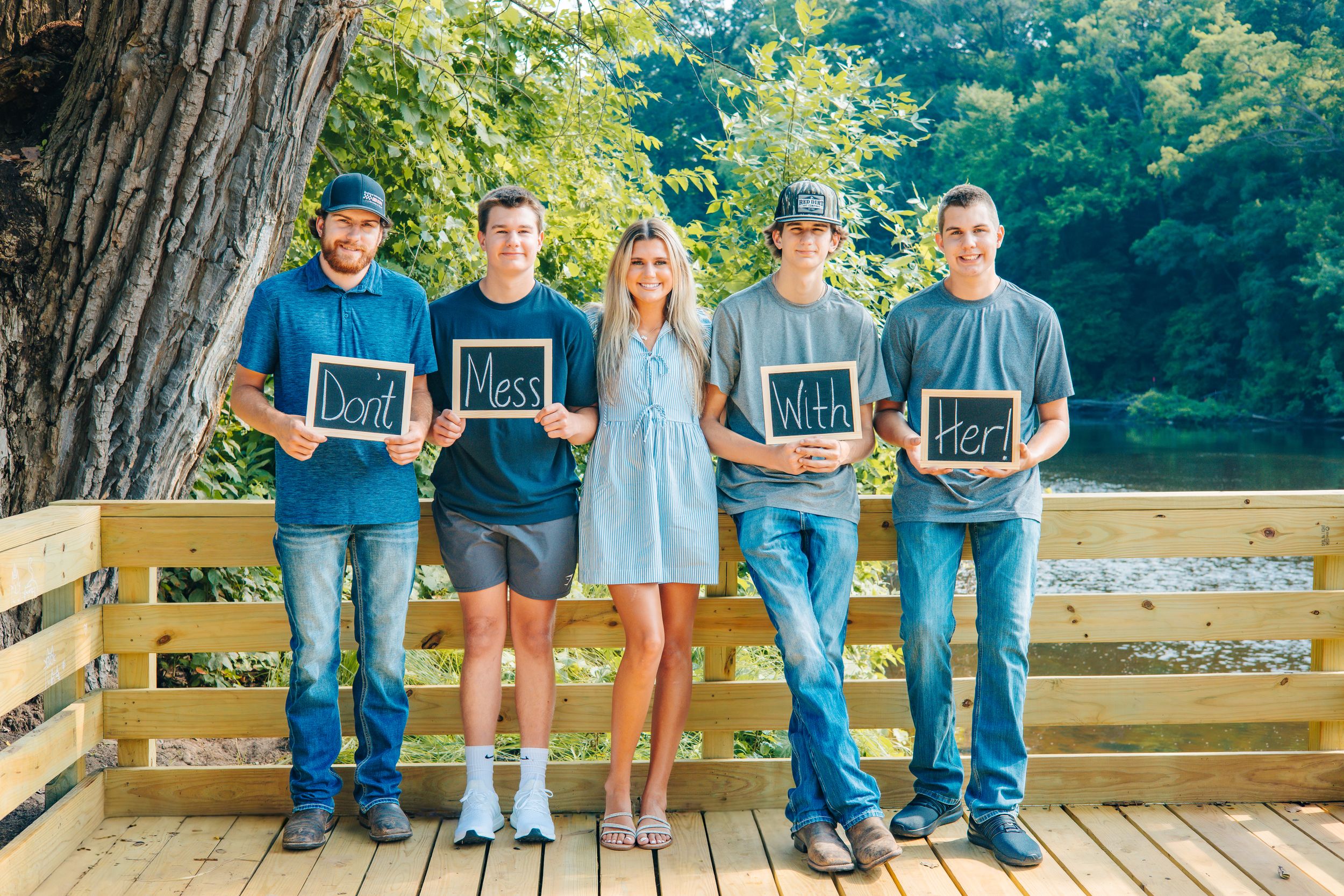 Five people stand on a wooden deck holding chalkboard signs reading Don't Mess With Her, posing outdoors near a tree.