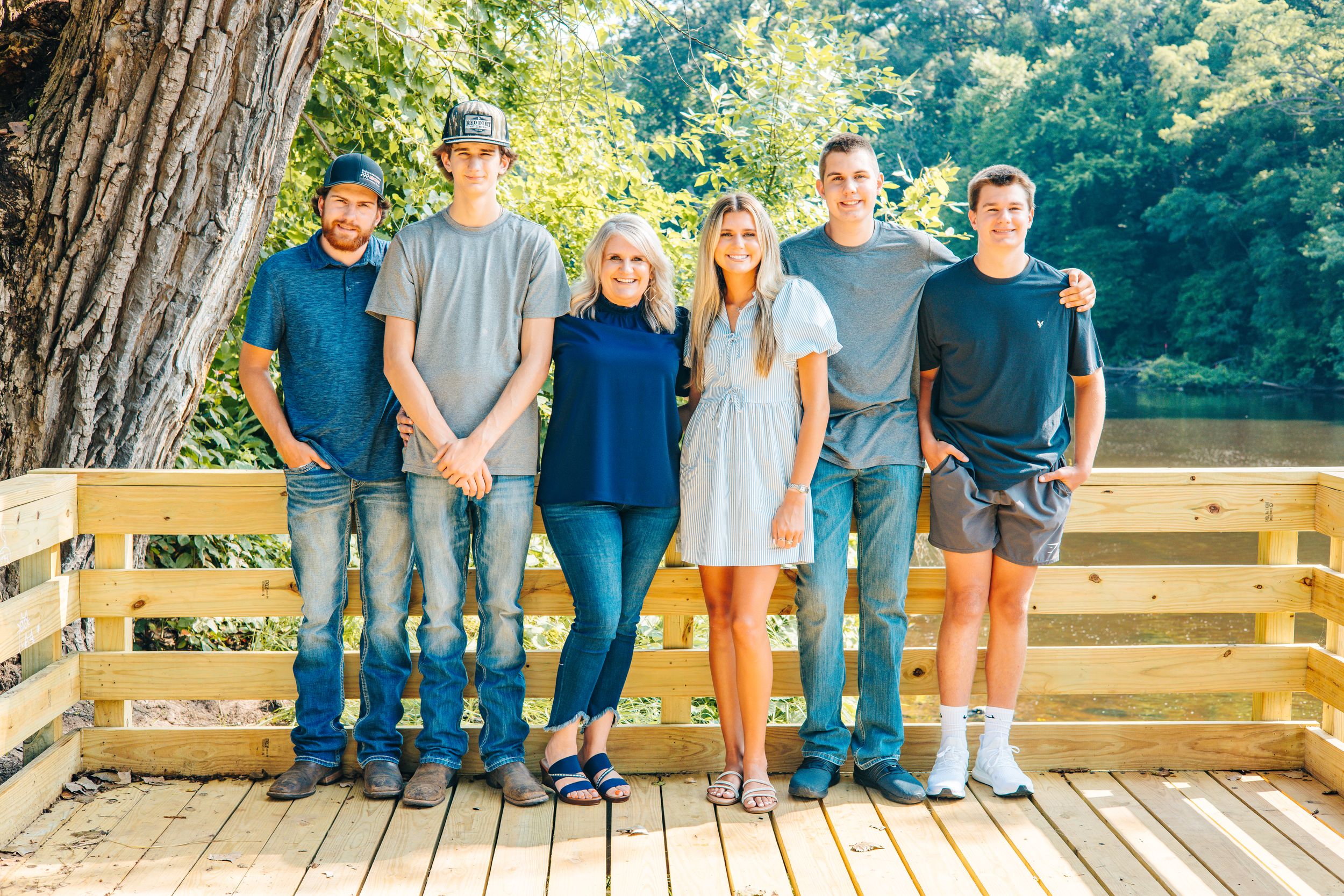 Family of six poses on a wooden deck surrounded by lush green trees near a serene lake.