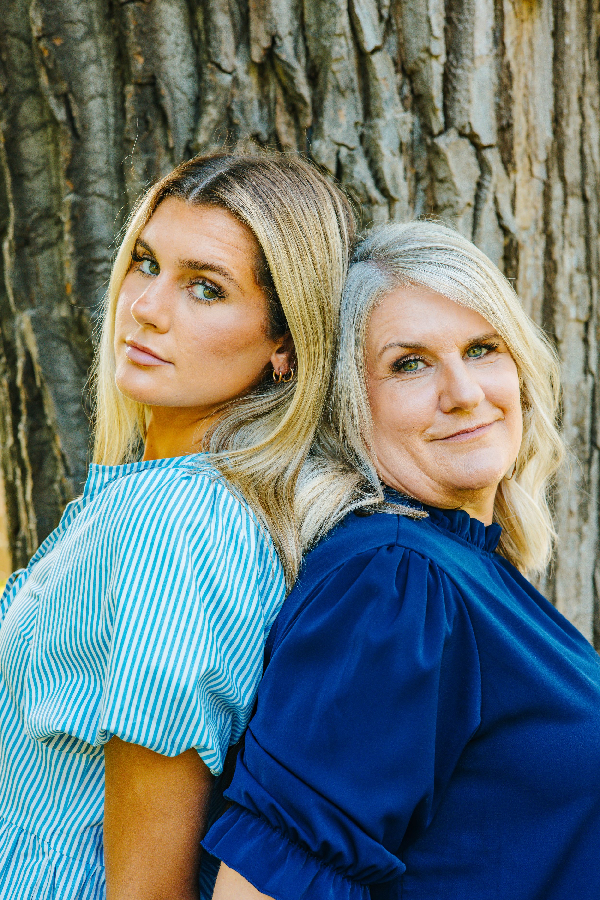 Two blonde women in blue outfits pose back-to-back against a tree, smiling over their shoulders.