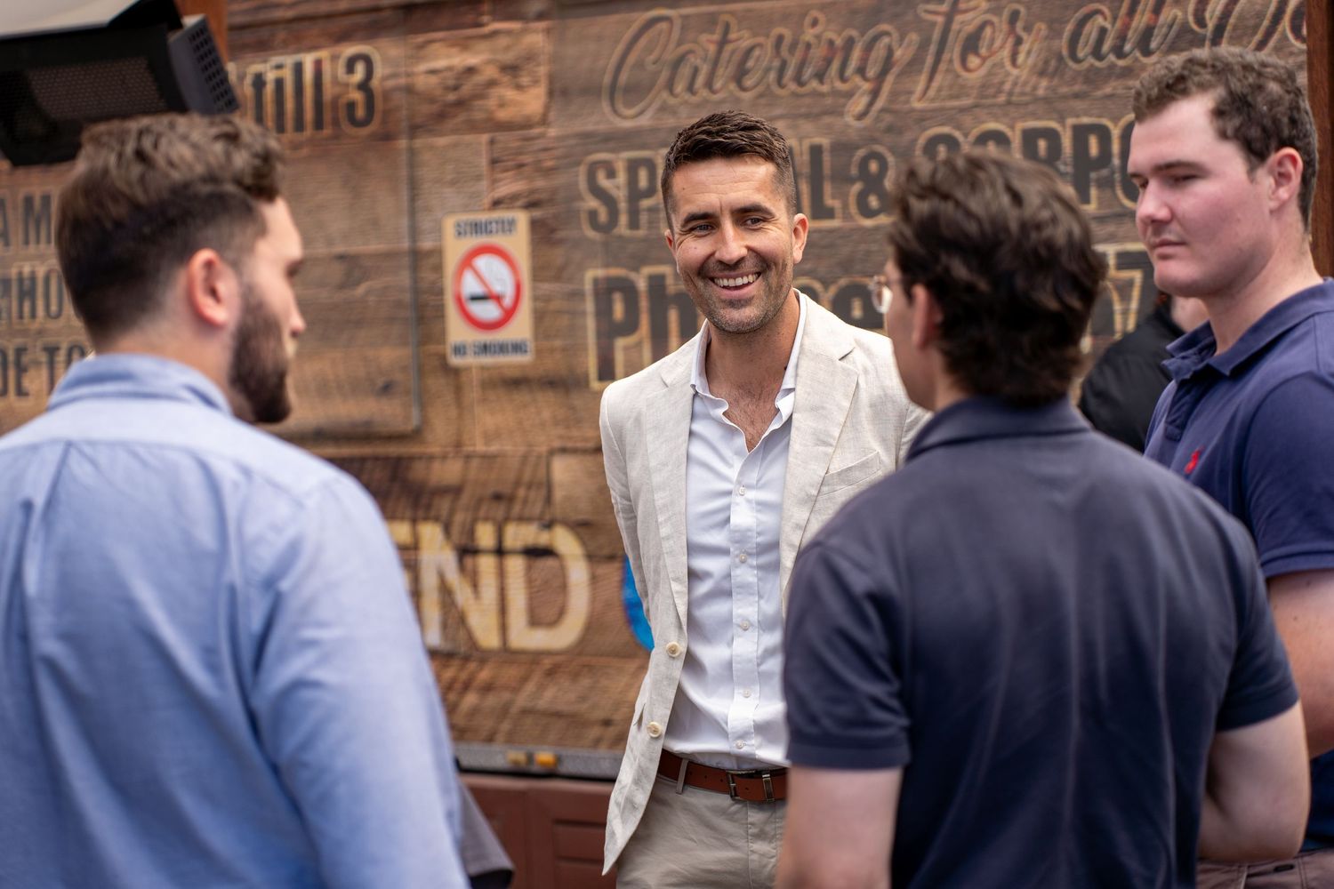 Smartly dressed man in beige suit smiling and engaging with group of young men at outdoor business event.