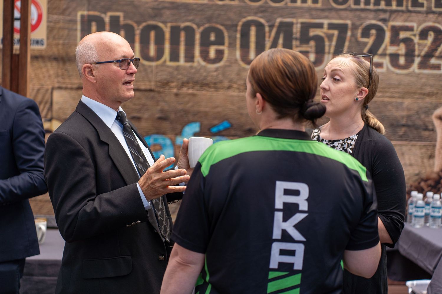 Business professionals in formal attire conversing with person in RKA sports jersey at outdoor corporate event.