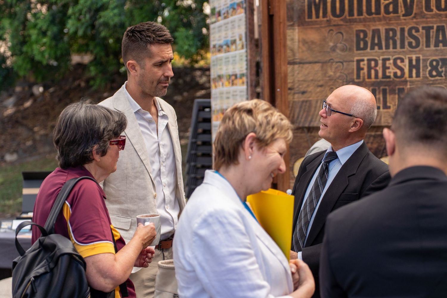 Group of five business professionals networking and conversing at outdoor corporate daytime event.