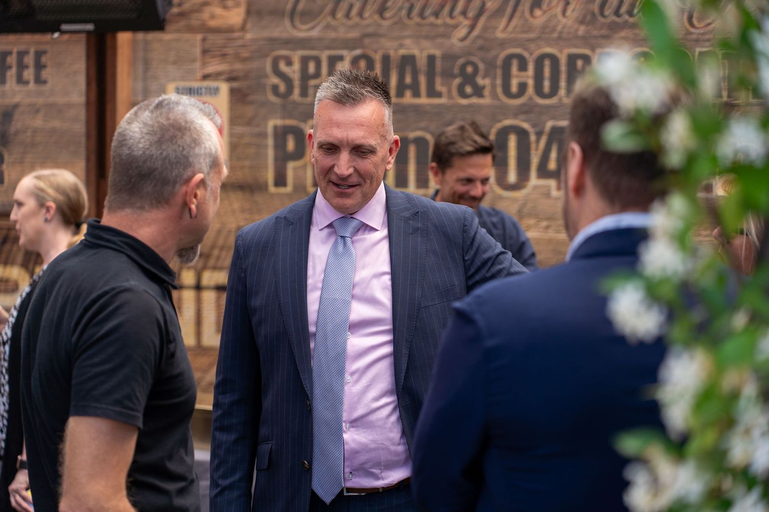 Confident businessman in pink shirt and navy suit smiling while networking at outdoor corporate event.