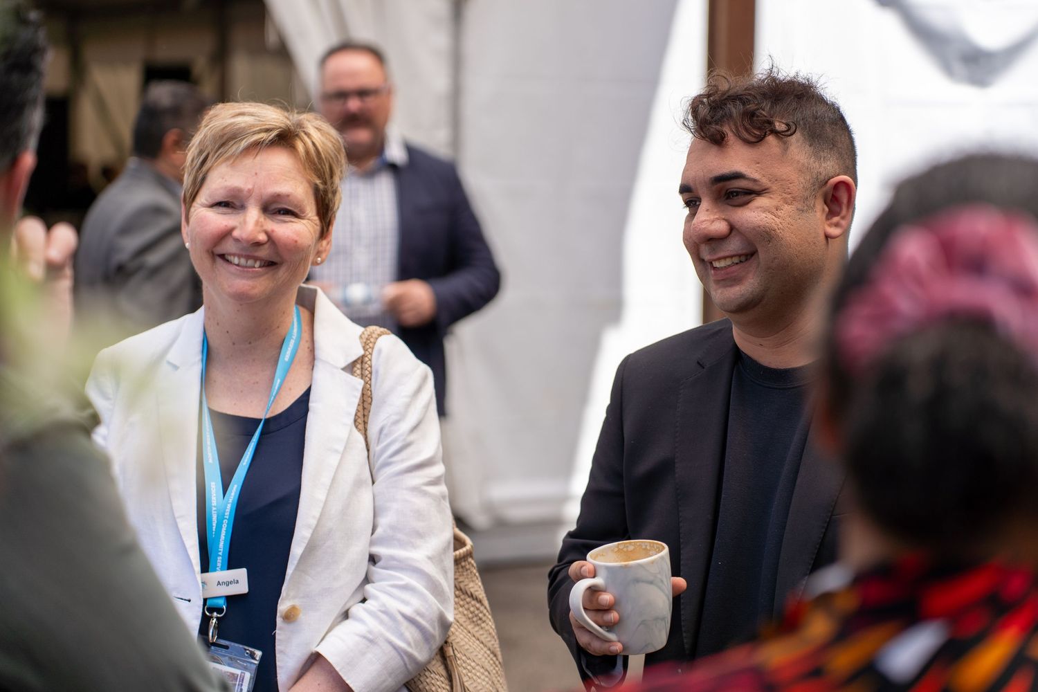 Smiling woman in white blazer and man in black jacket networking with coffee cups at business conference.