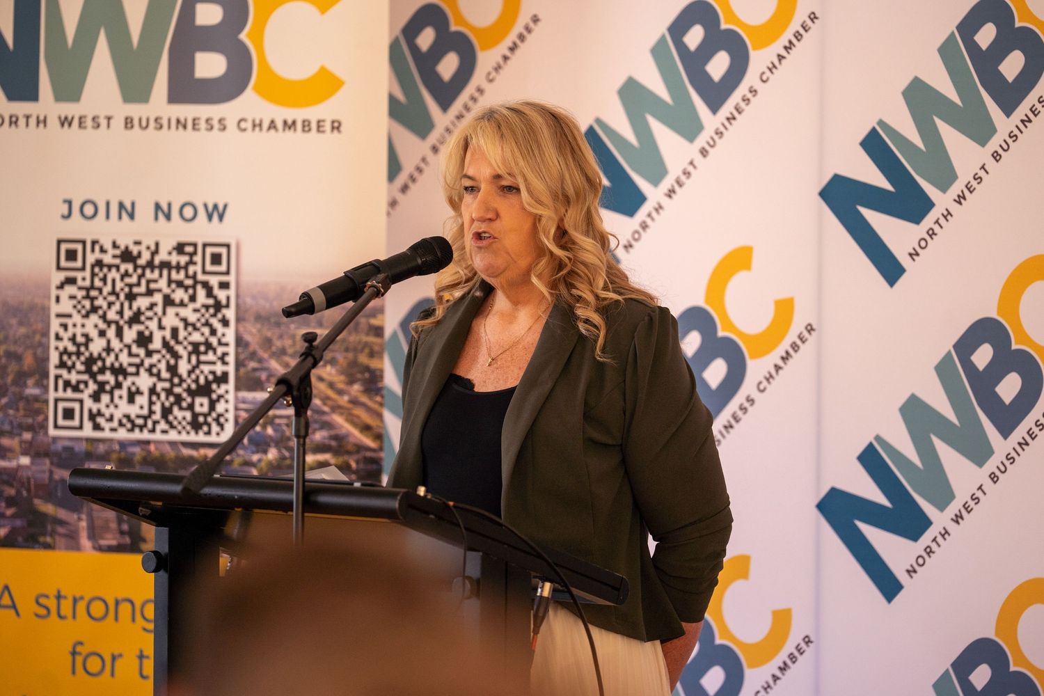 Woman presenting at a North West Business Chamber NWBC podium with QR code banner.
