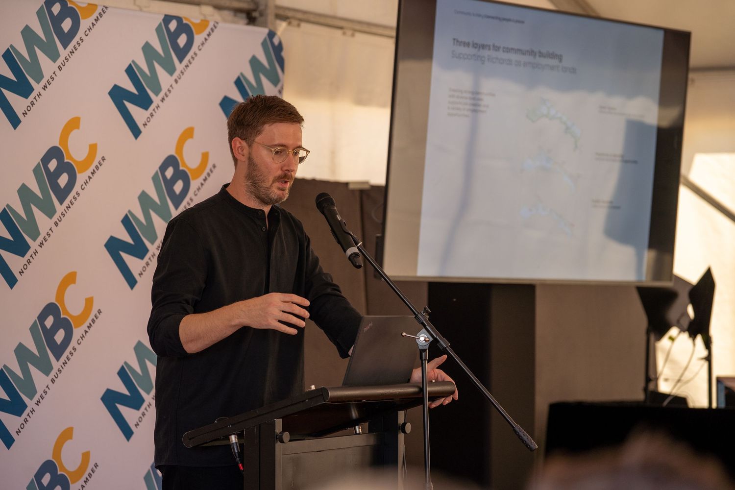 Man in black presenting at NWBC business event podium with projection screen displaying data behind him.