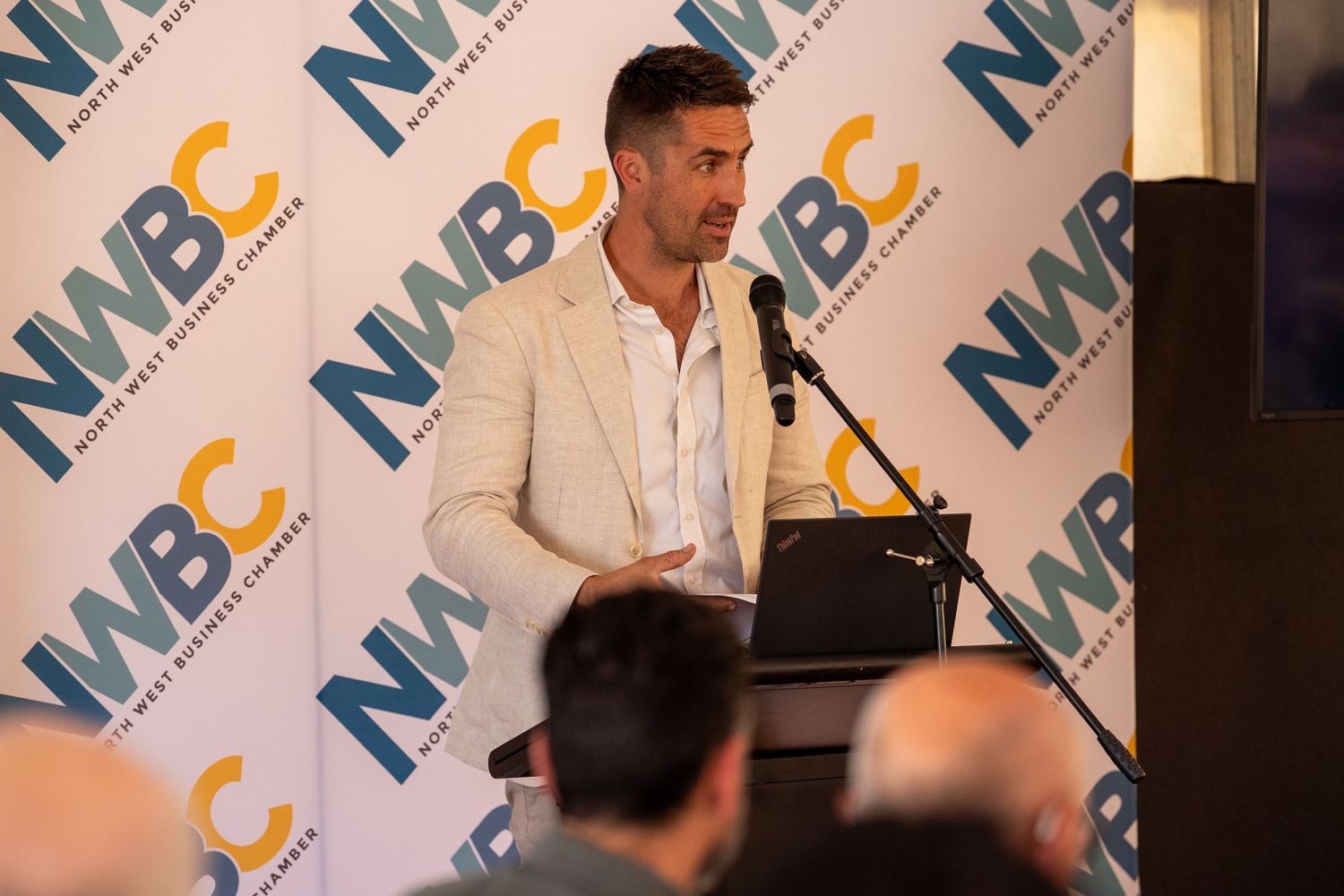 Man in beige blazer speaking at NWBC business chamber podium event before seated audience.