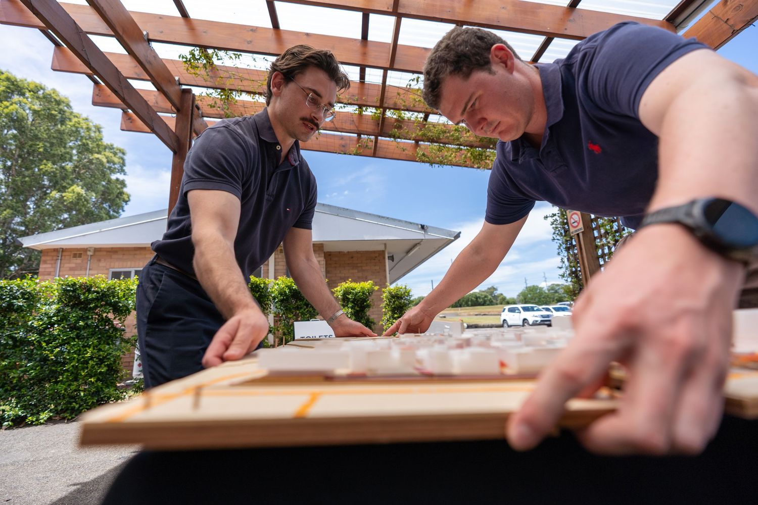 Two men collaborating over large architectural map on outdoor table under wooden pergola.