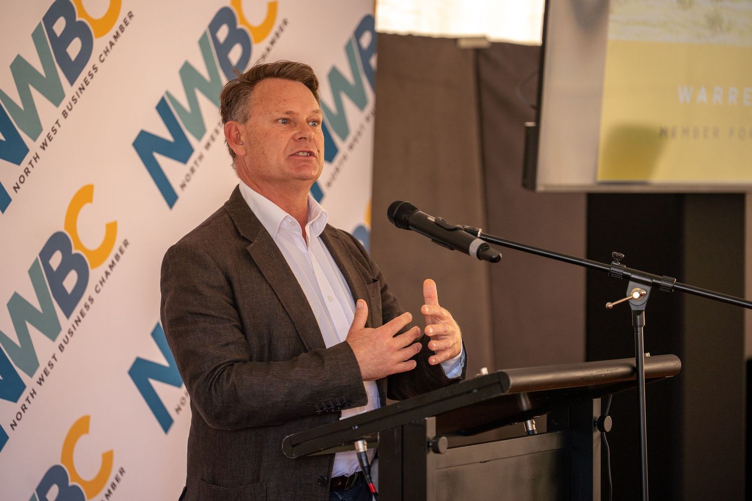 Tall man in grey suit speaking at NWBC business chamber podium with microphone.