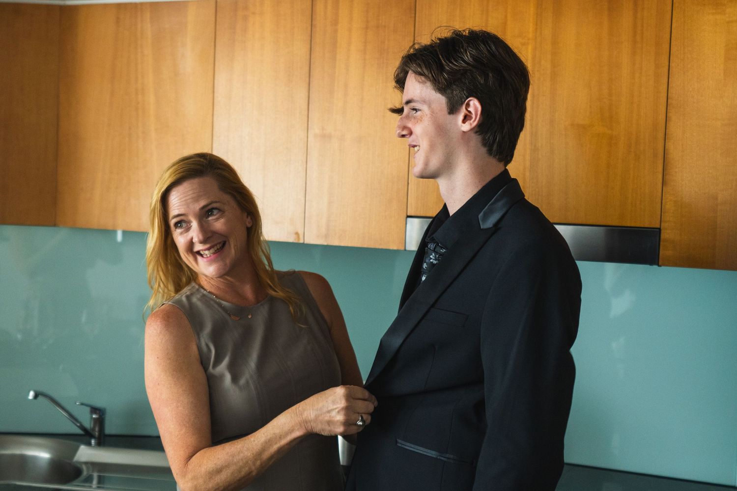 Woman smiling while adjusting a young mans suit jacket in a modern kitchen.