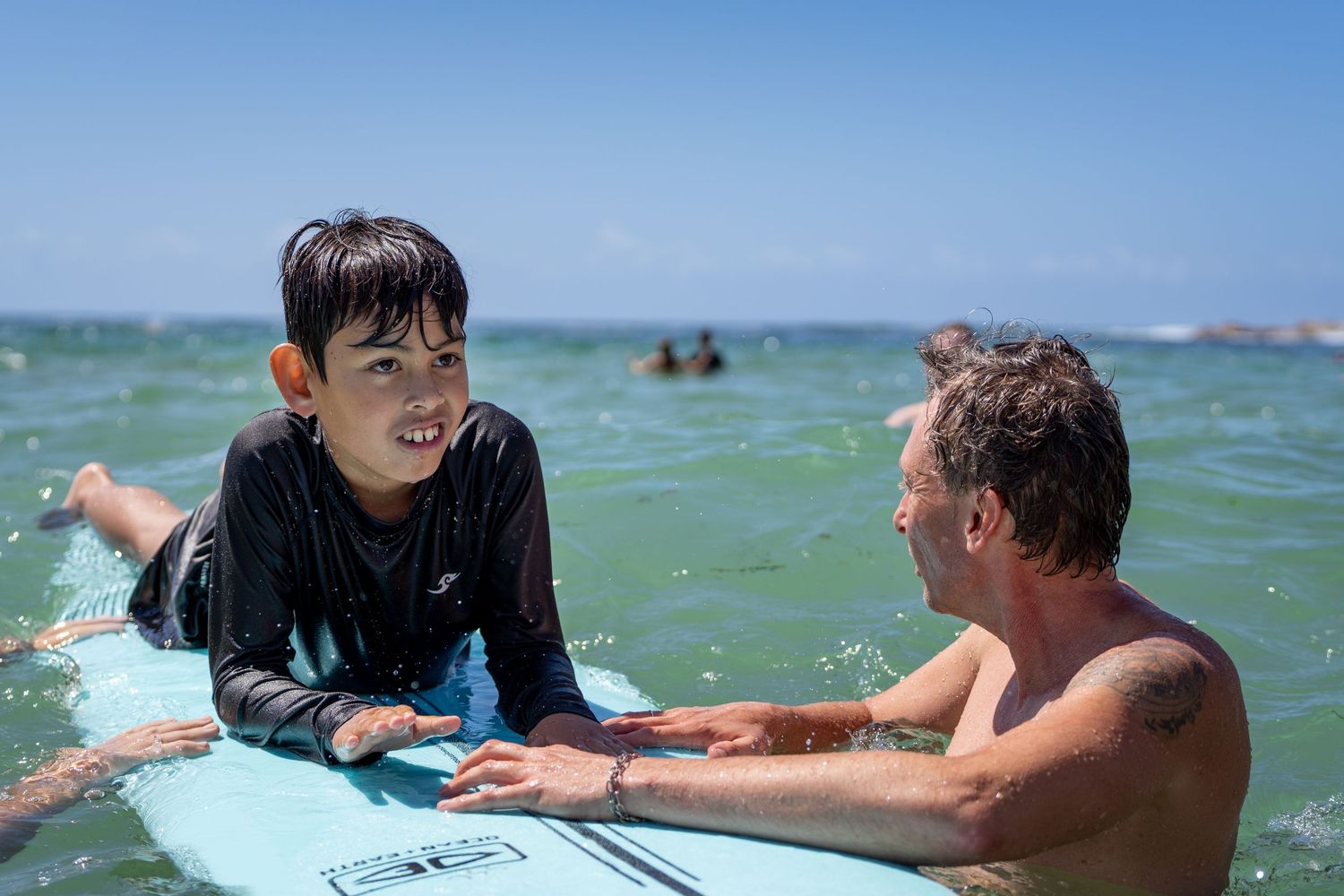 A young boy lies on a surfboard in the ocean while an adult surf instructor guides him during a lesson.