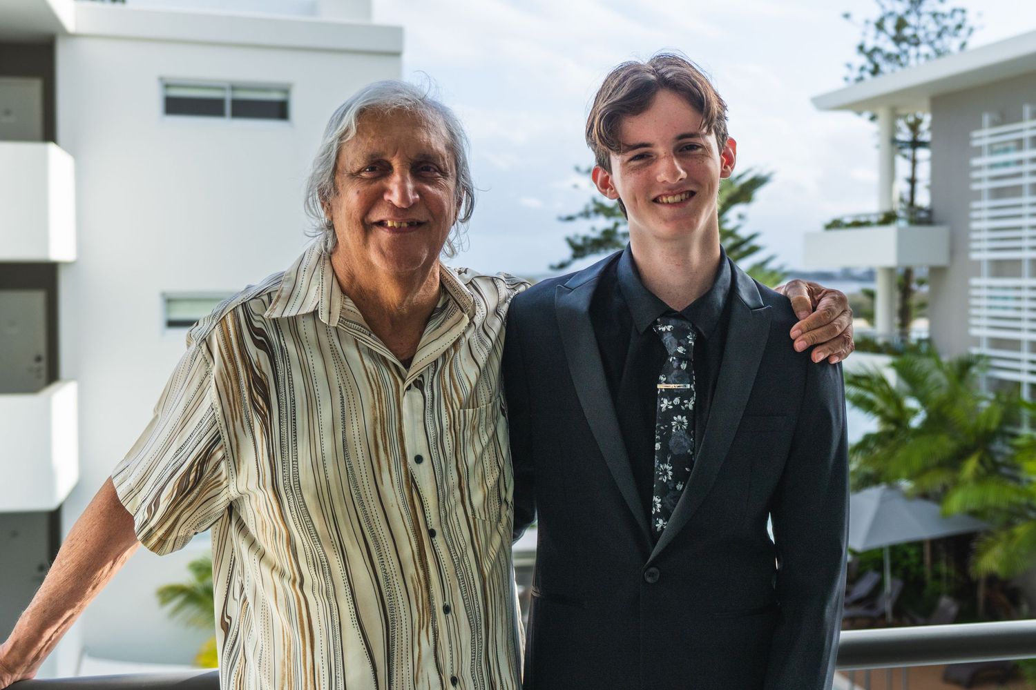 Elderly man and tall teenage boy in a suit smiling together on an outdoor balcony.