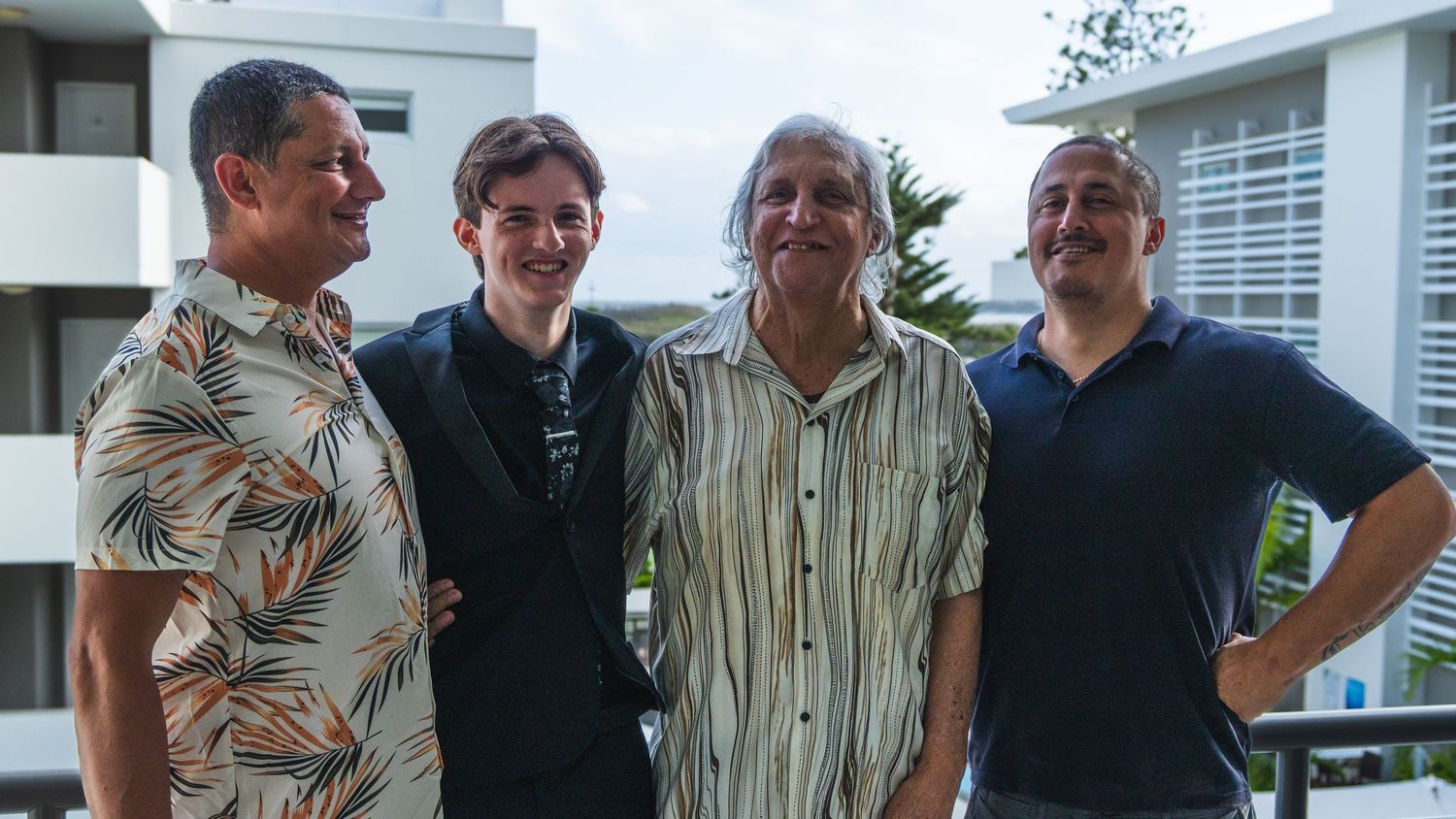 Four men of varying ages smiling together for a group photo on an outdoor balcony.