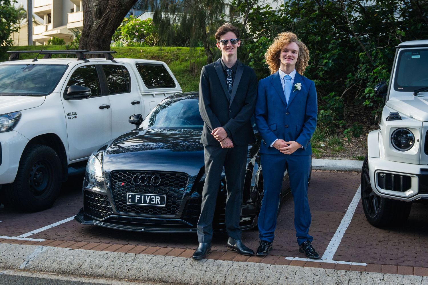 Two teenage boys dressed in formal suits standing in front of cars in a parking lot.