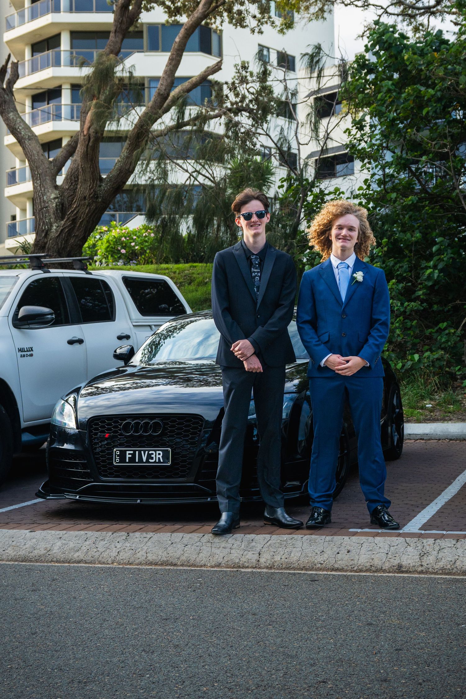 Two formally dressed teenage boys pose in front of a black Audi with FIV3R plates at a school formal.
