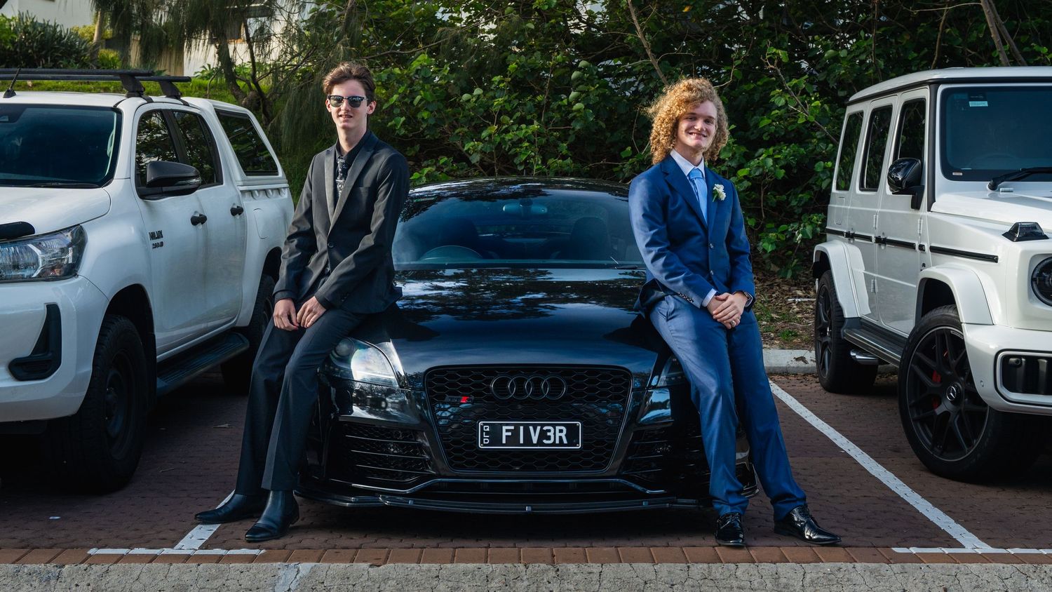 Two formally dressed teens lean against a black Audi FIV3R at a school formal photoshoot.