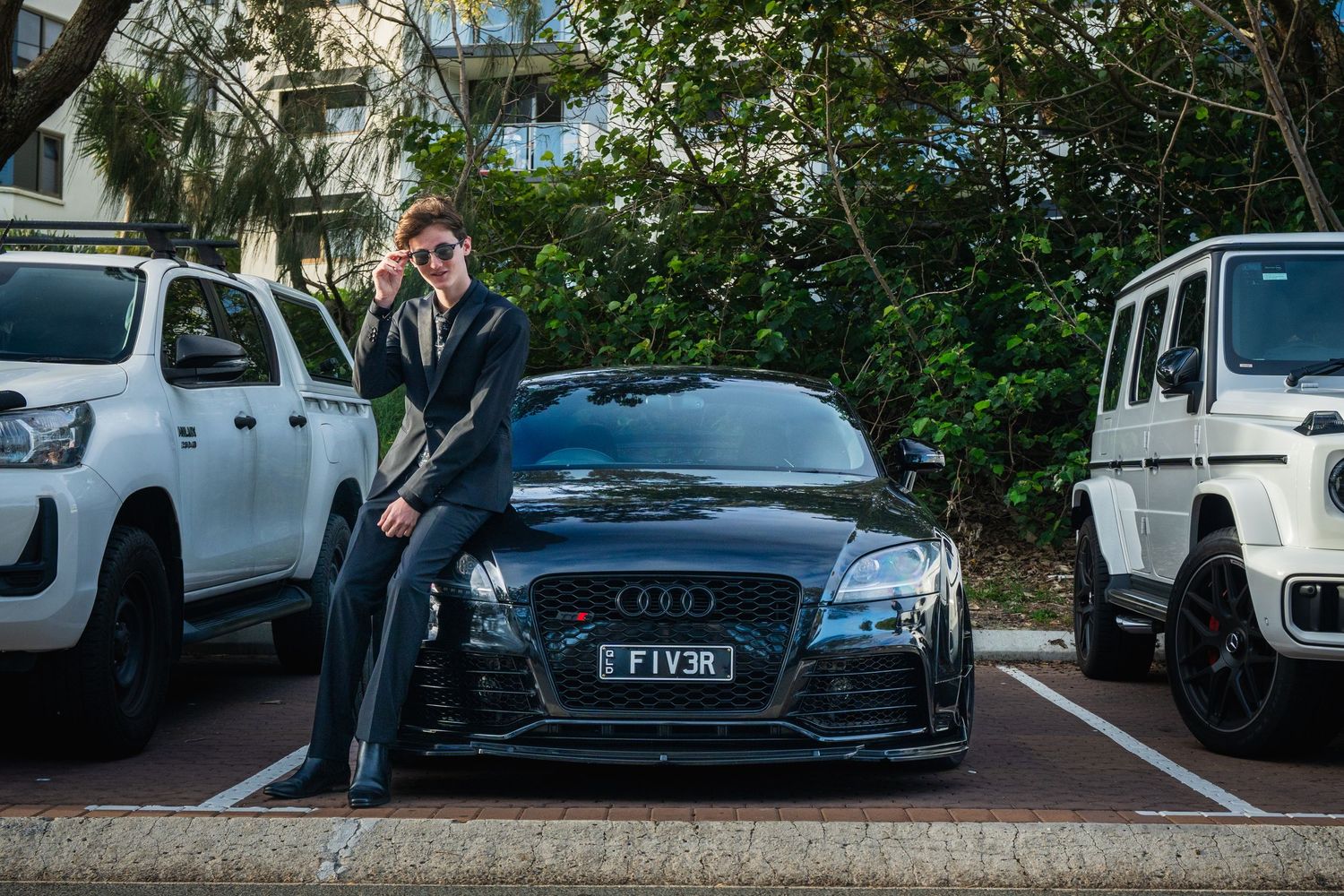 A young man in a dark suit poses beside a black Audi TT with custom FIV3R number plate.