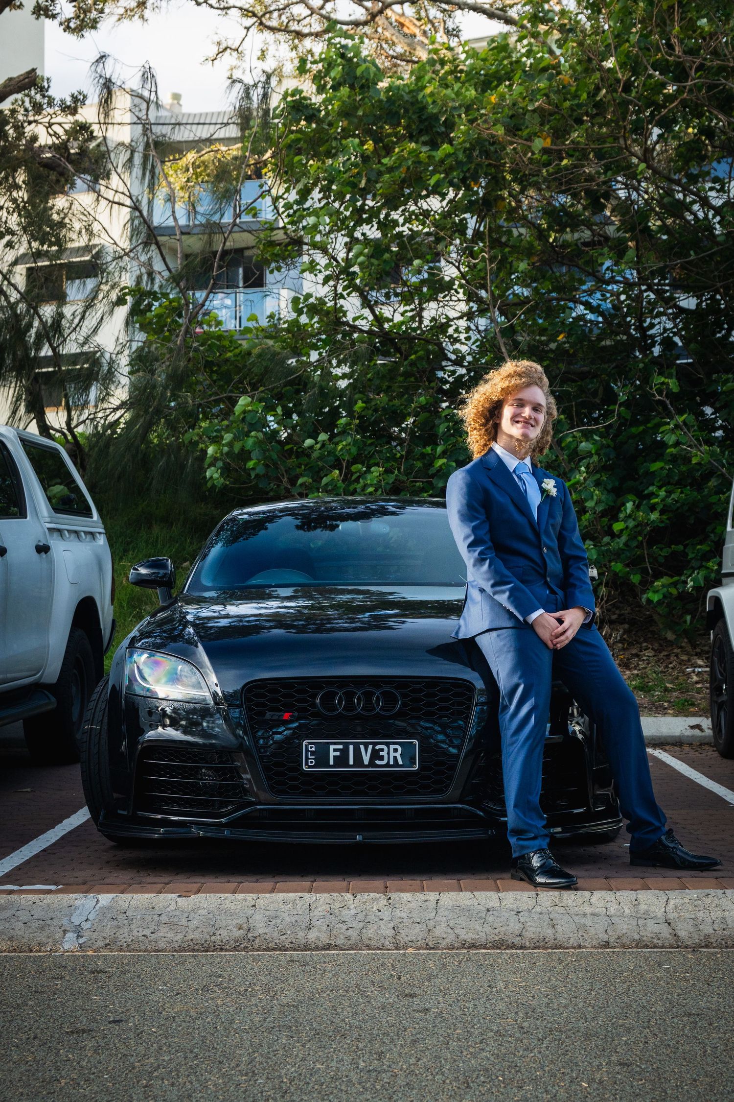 A curly-haired teen in a blue suit sits on a black Audi at a school formal event.