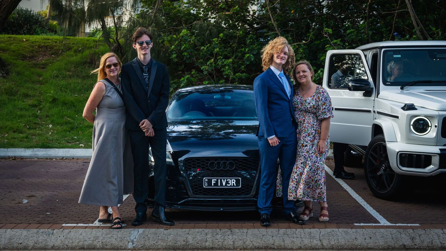 Two formally dressed teens with their parents pose beside a black Audi FIV3R before school formal.