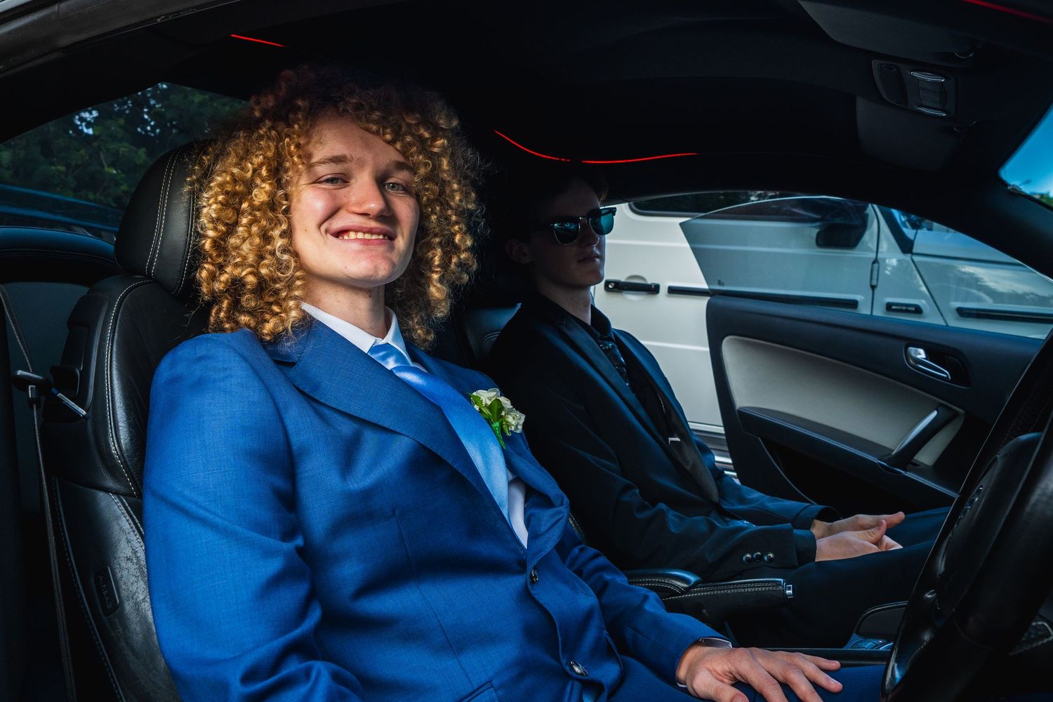 Two formally dressed teenage boys smile inside a black car heading to school formal.