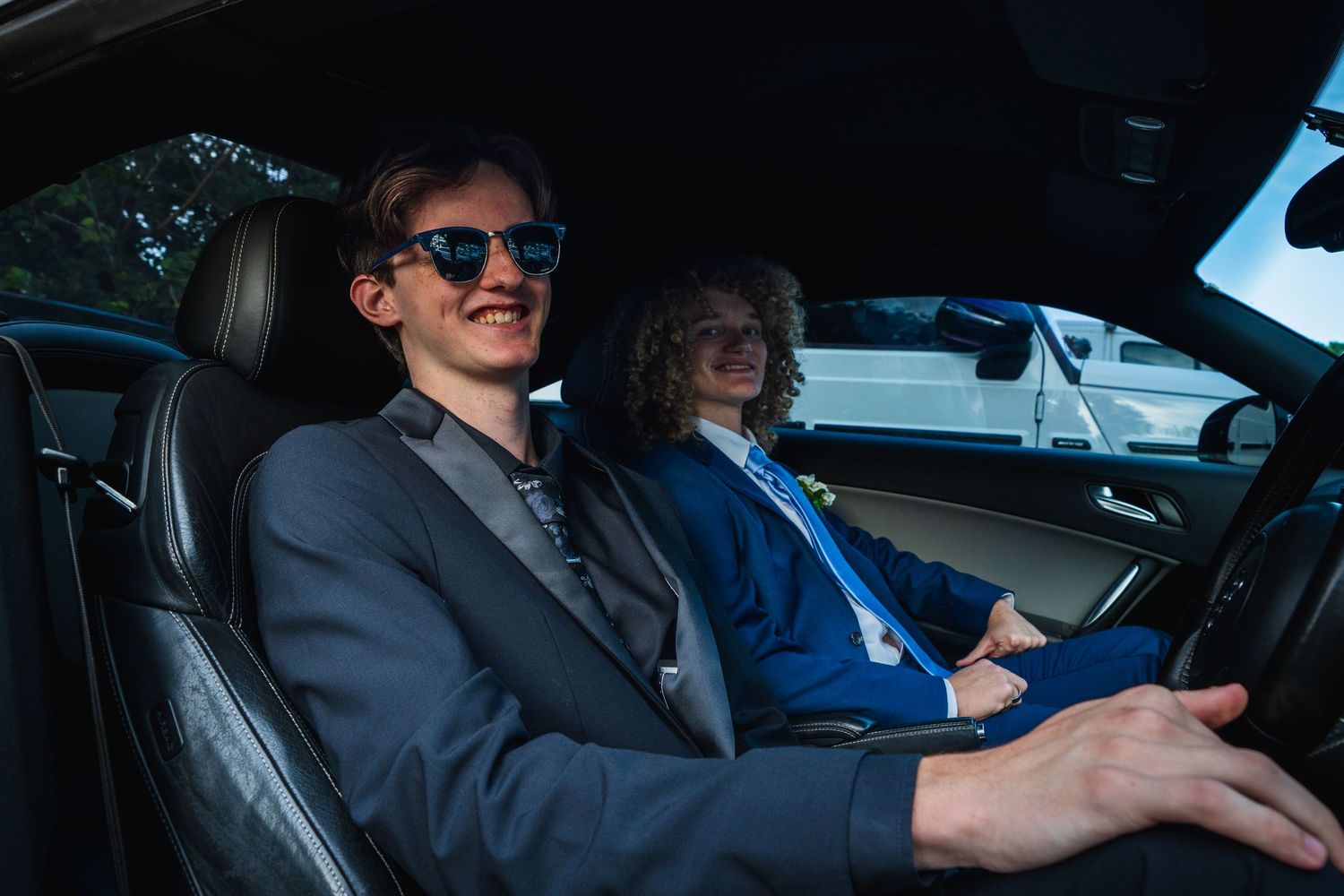 Two smartly dressed teenage boys sit in a black car driving to their school formal.