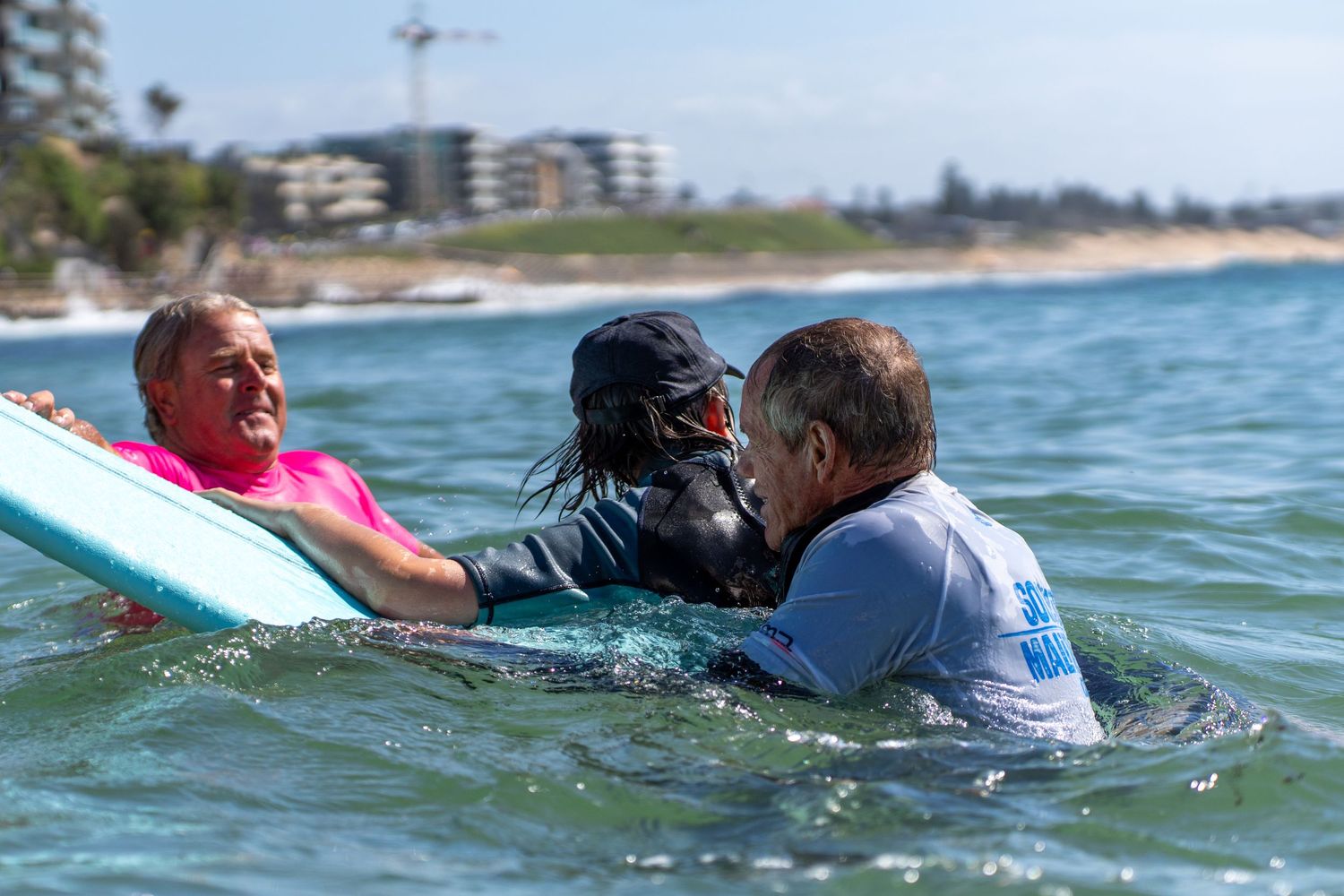 Two surf instructors assist a child in a wetsuit on a surfboard in the ocean near a coastal town.