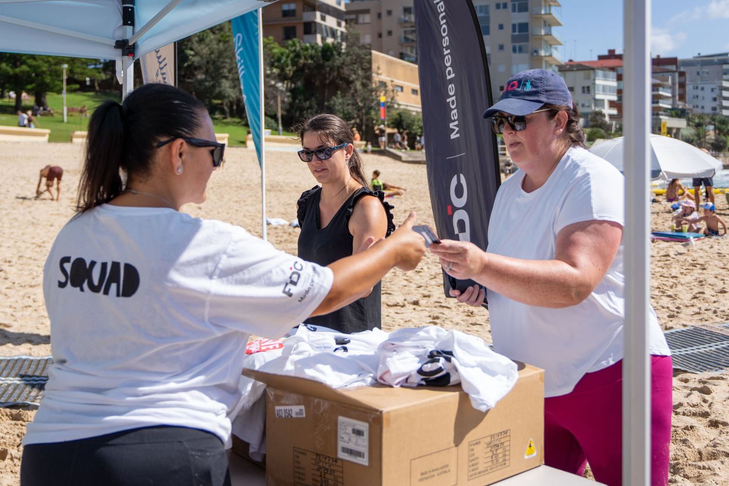 Three women exchange items at a beach event tent on a sunny day at a sandy beach.