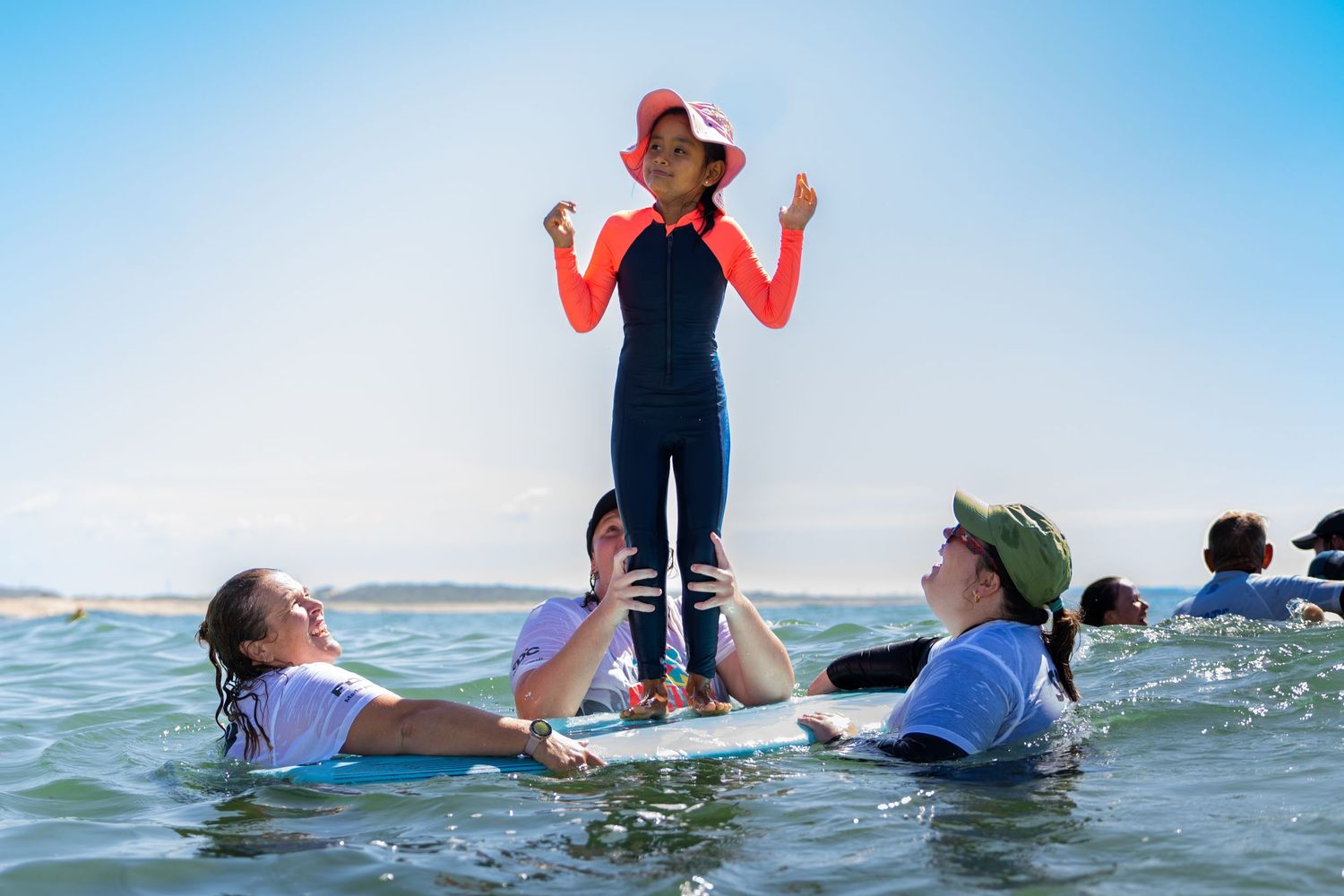 A child in a red hat stands triumphantly on a surfboard while two instructors support her in the water.