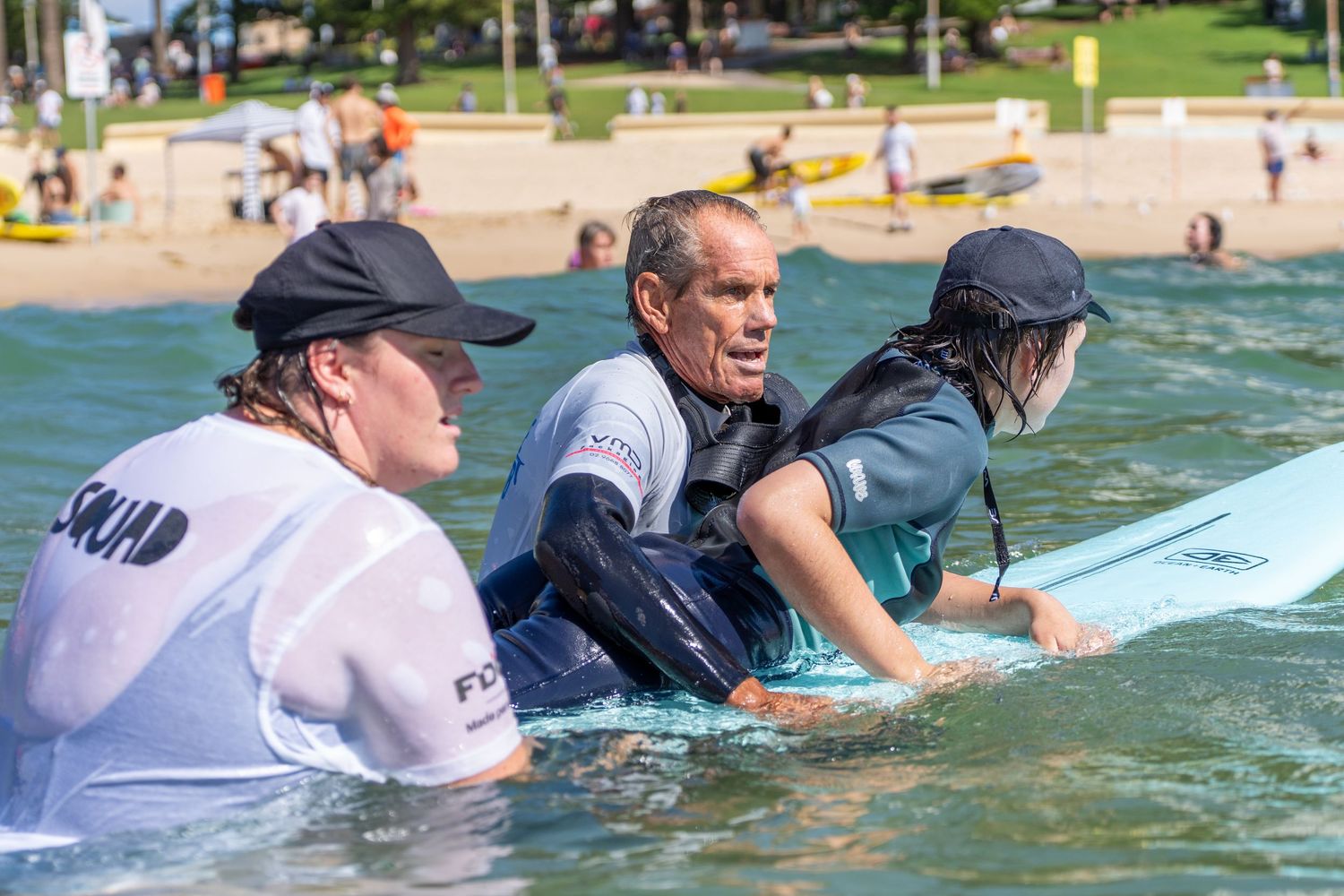 Two surf instructors wearing Squad shirts help a participant onto a surfboard in shallow ocean water.