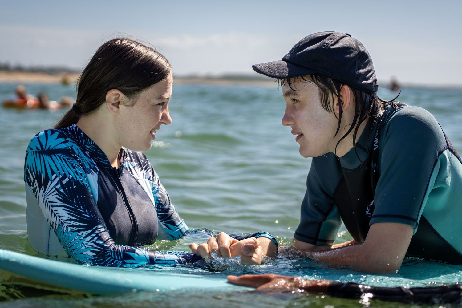 Two young women in wetsuits share a moment together while holding onto a surfboard in the ocean.