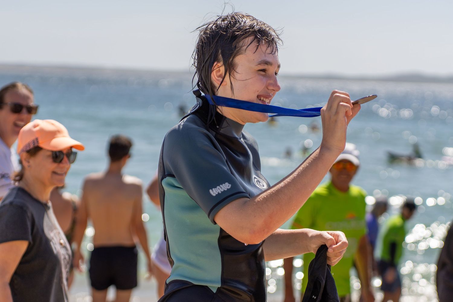A young person in a wetsuit adjusts their chin strap on a sunny beach with people in the background.